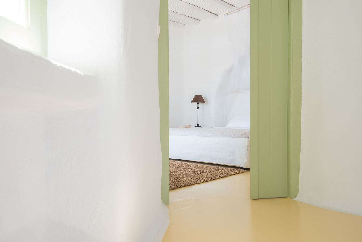 Minimalist white hallway with green doorframe leading to sunlit bedroom with natural jute rug.