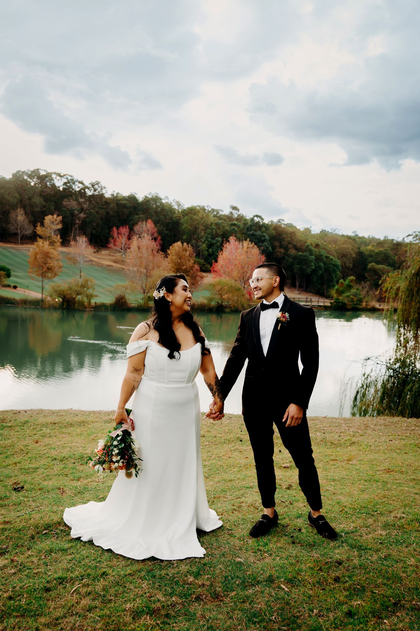 Bride and groom holding hands by a serene lake with autumn foliage in the background.