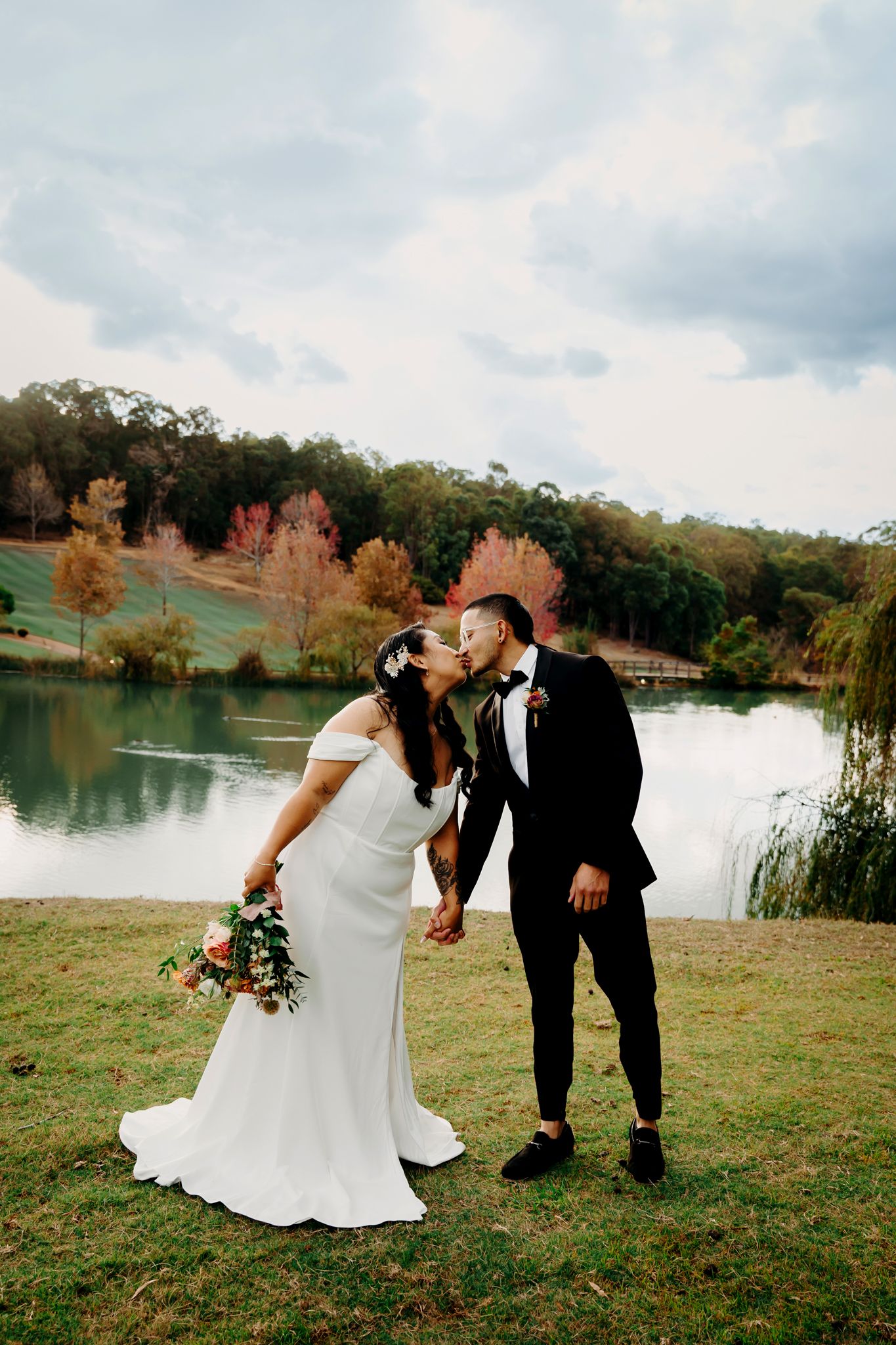 Newlyweds embrace by a serene lake with autumn trees in the background.
