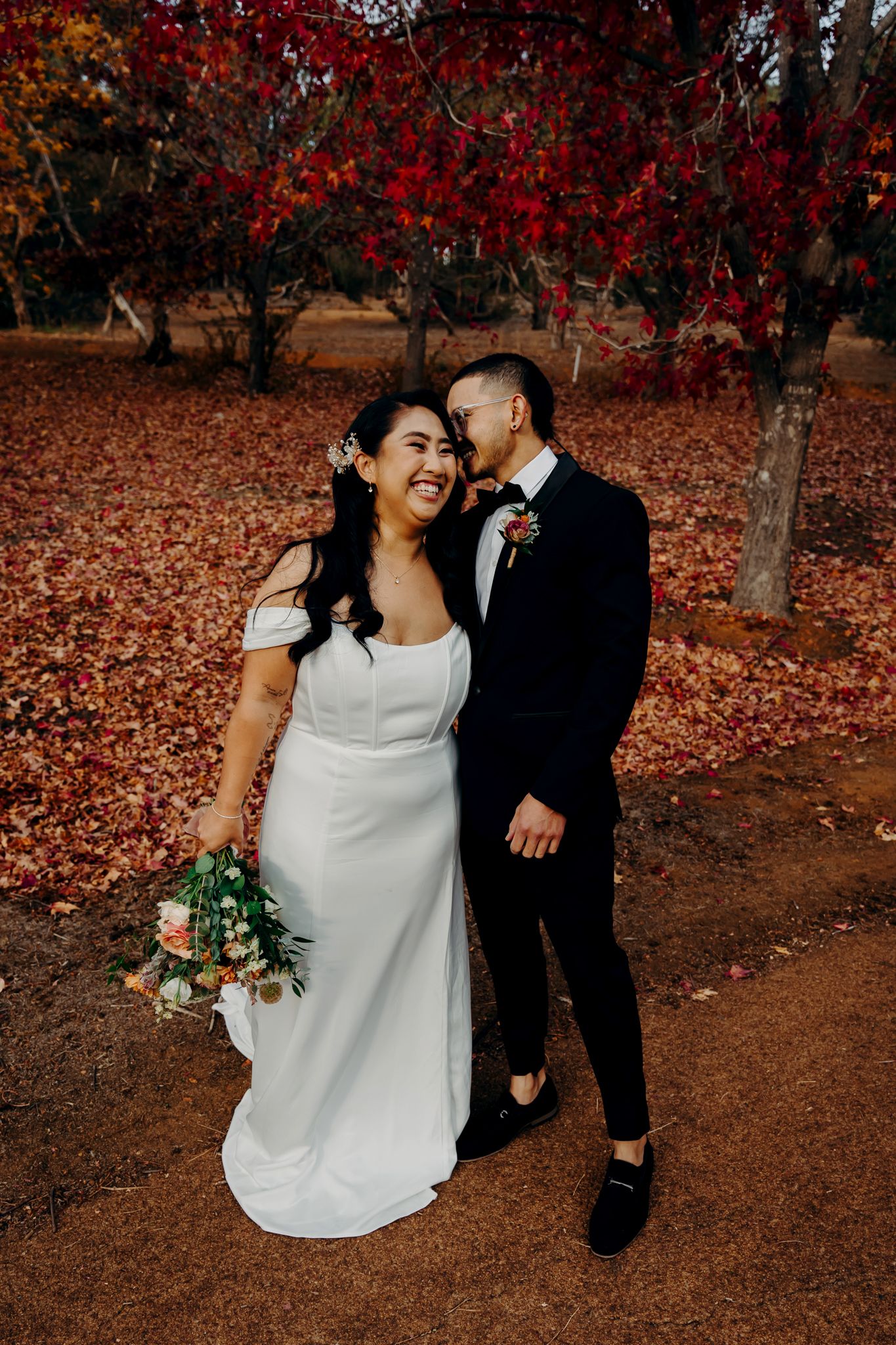 Wedding portrait under vibrant red autumn trees with fallen leaves.