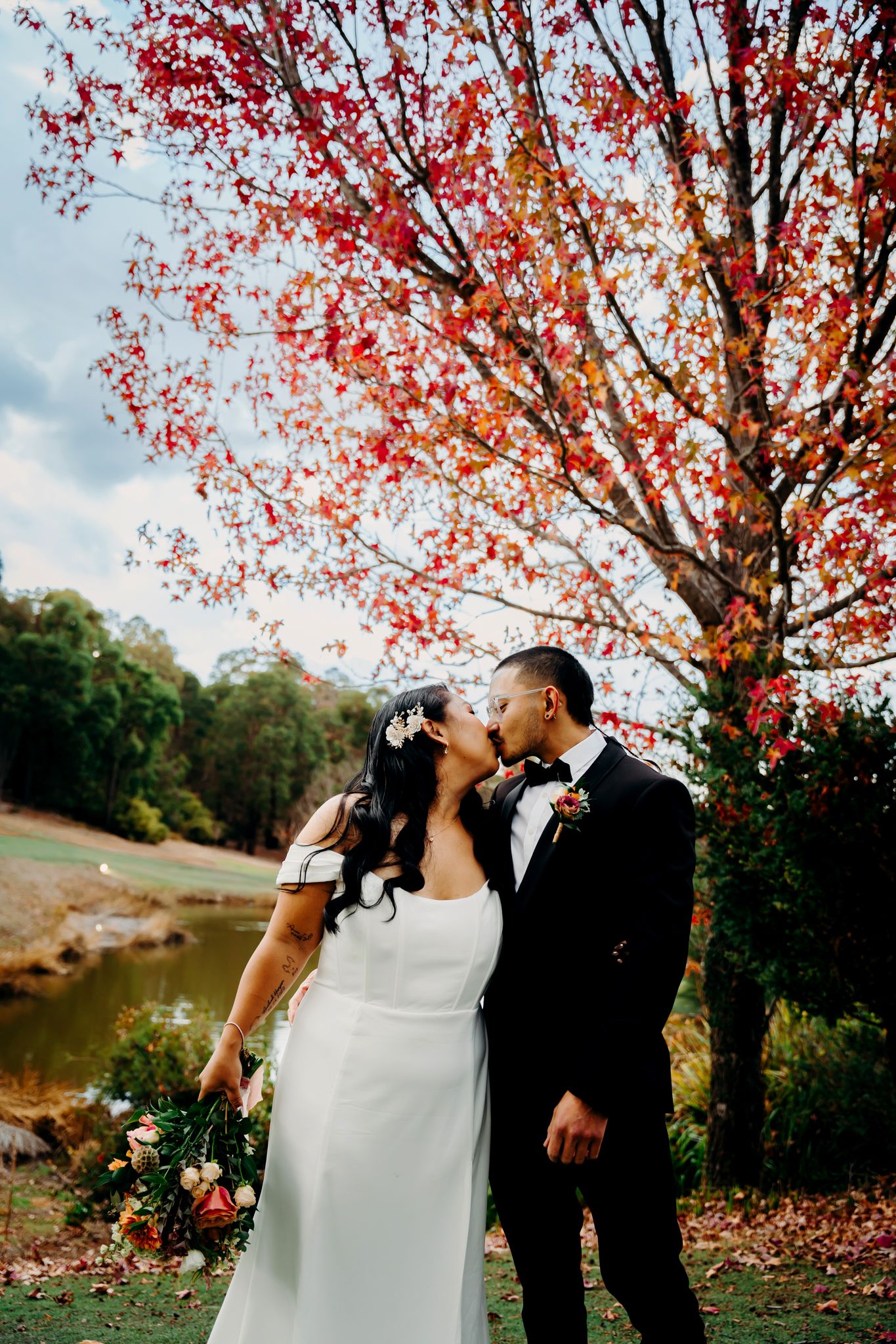 Romantic wedding moment under a bright red autumn tree near a pond.
