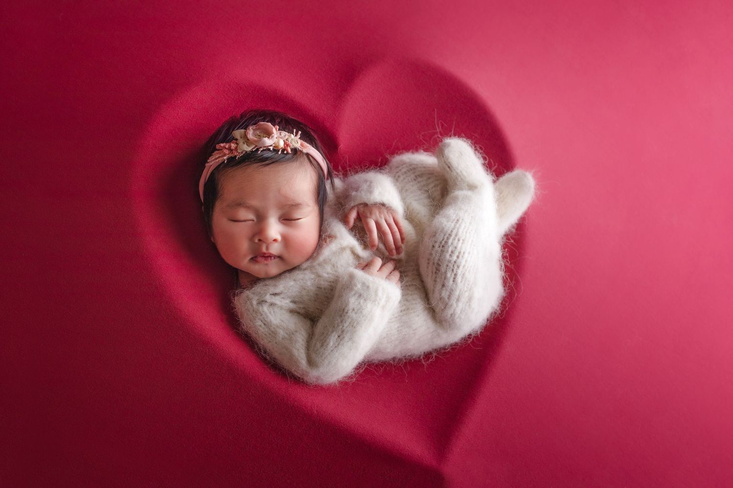 Sleeping newborn in white knit wrap inside heart-shaped red backdrop.