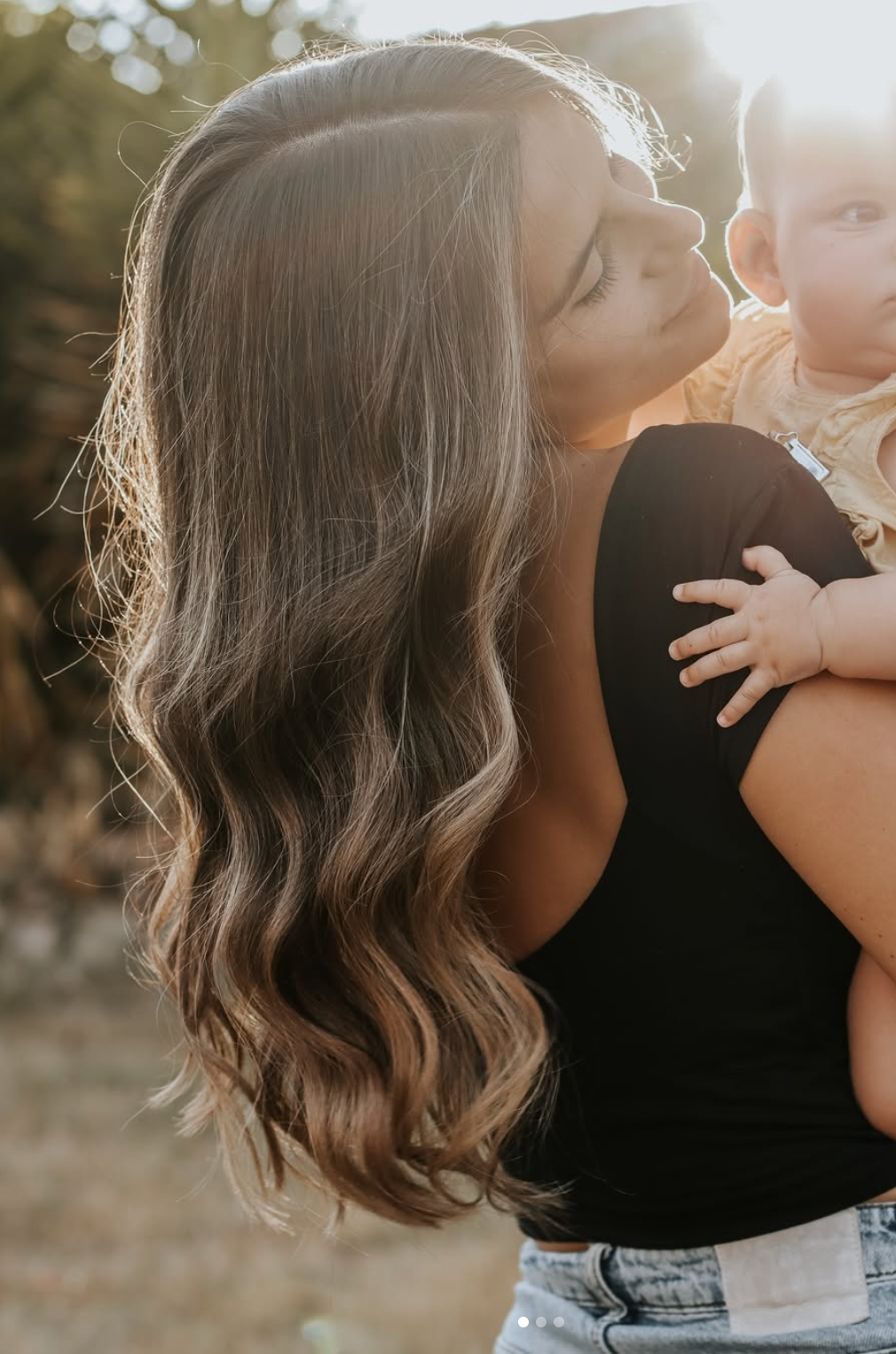 A mother with long wavy brunette hair kisses her baby in golden sunlight outdoors.