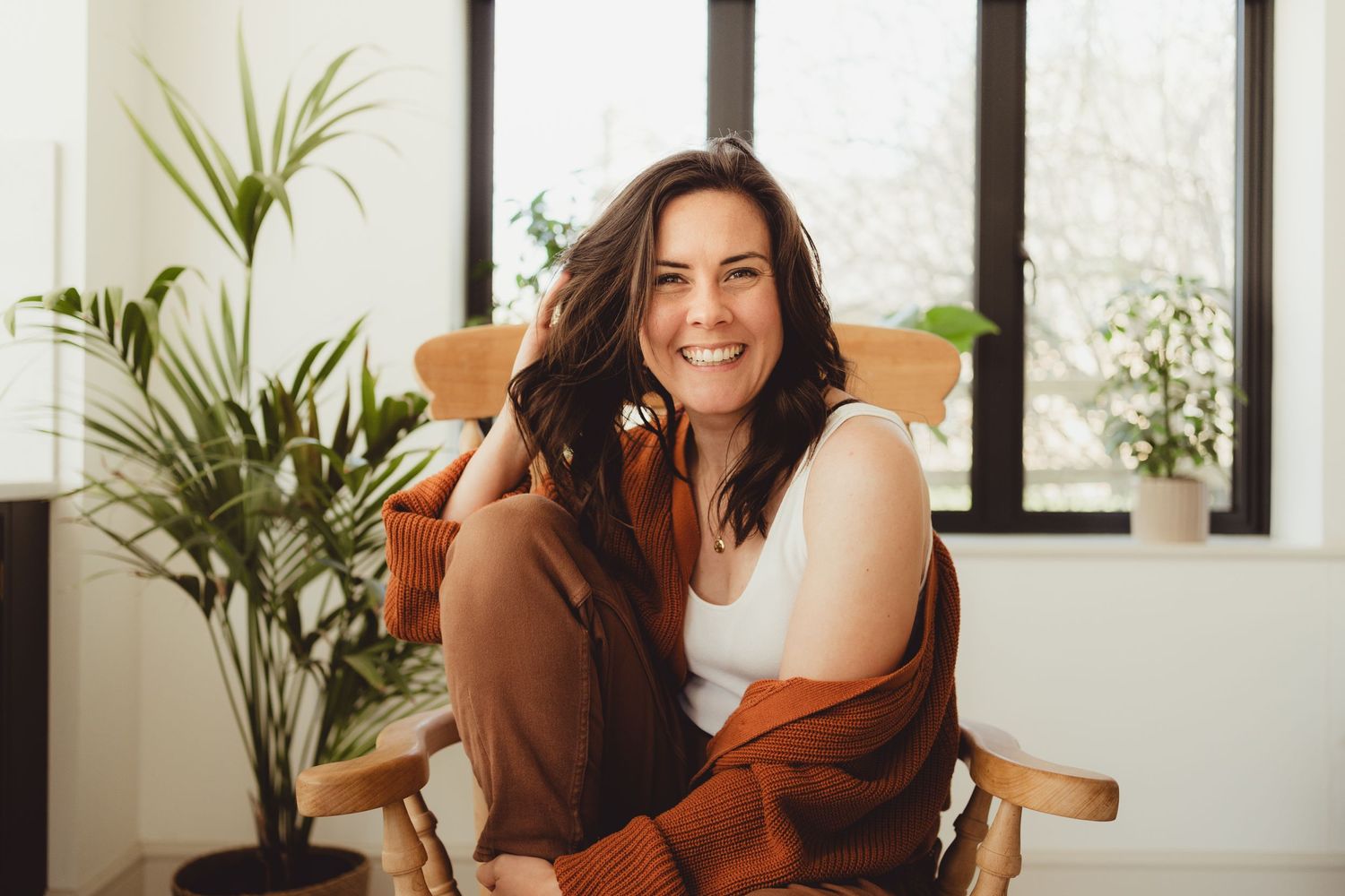 Warm toned indoor portrait of someone sitting comfortably in wooden chair with houseplants and natural lighting.
