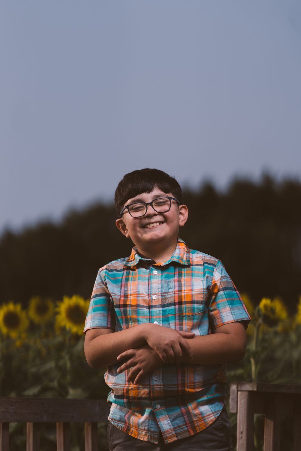 Child in plaid shirt and glasses smiles while leaning on wooden fence near sunflower field.