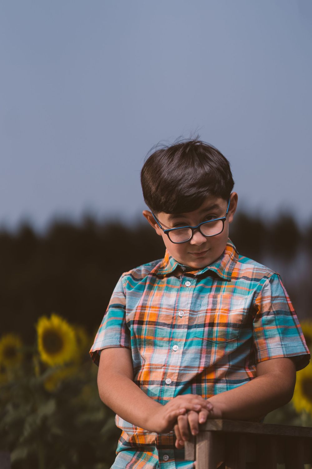 Child in glasses and plaid shirt laughs while resting arms on wooden fence.