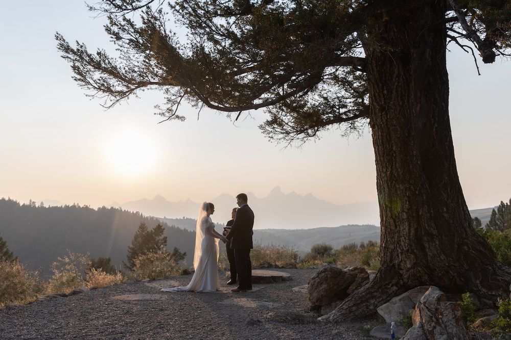 Couple exchanging vows at sunset under a large tree on a mountain overlook with misty peaks in the background.