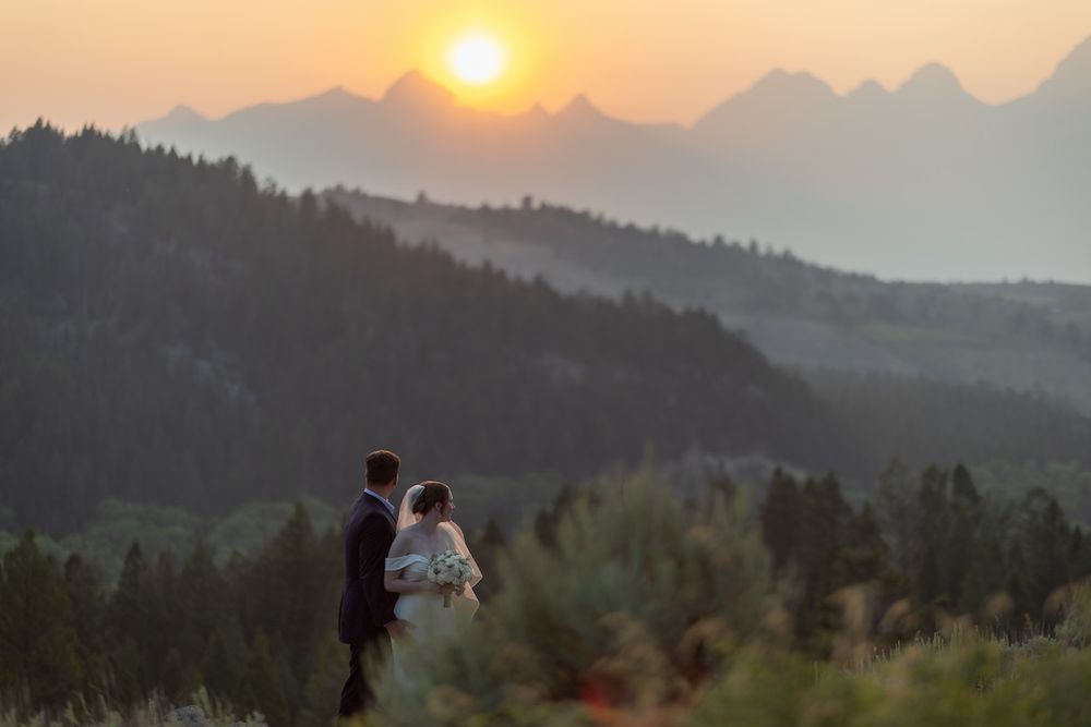 Couple stands together watching a stunning mountain sunset with layered peaks and forest in the background.