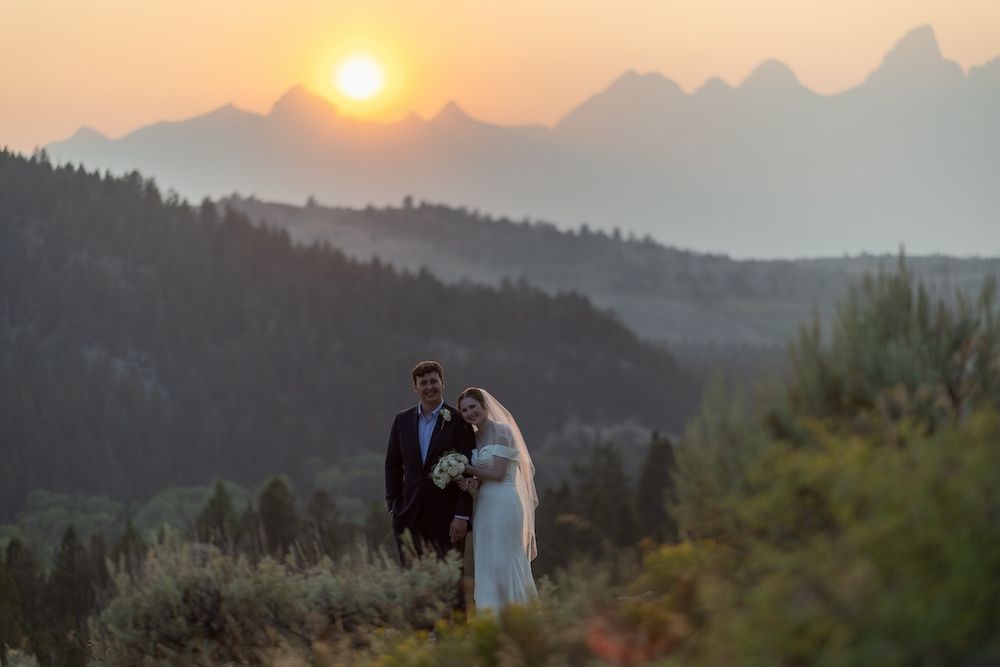 Bride and groom pose at sunset with Grand Teton mountain range silhouette in background during outdoor wedding.