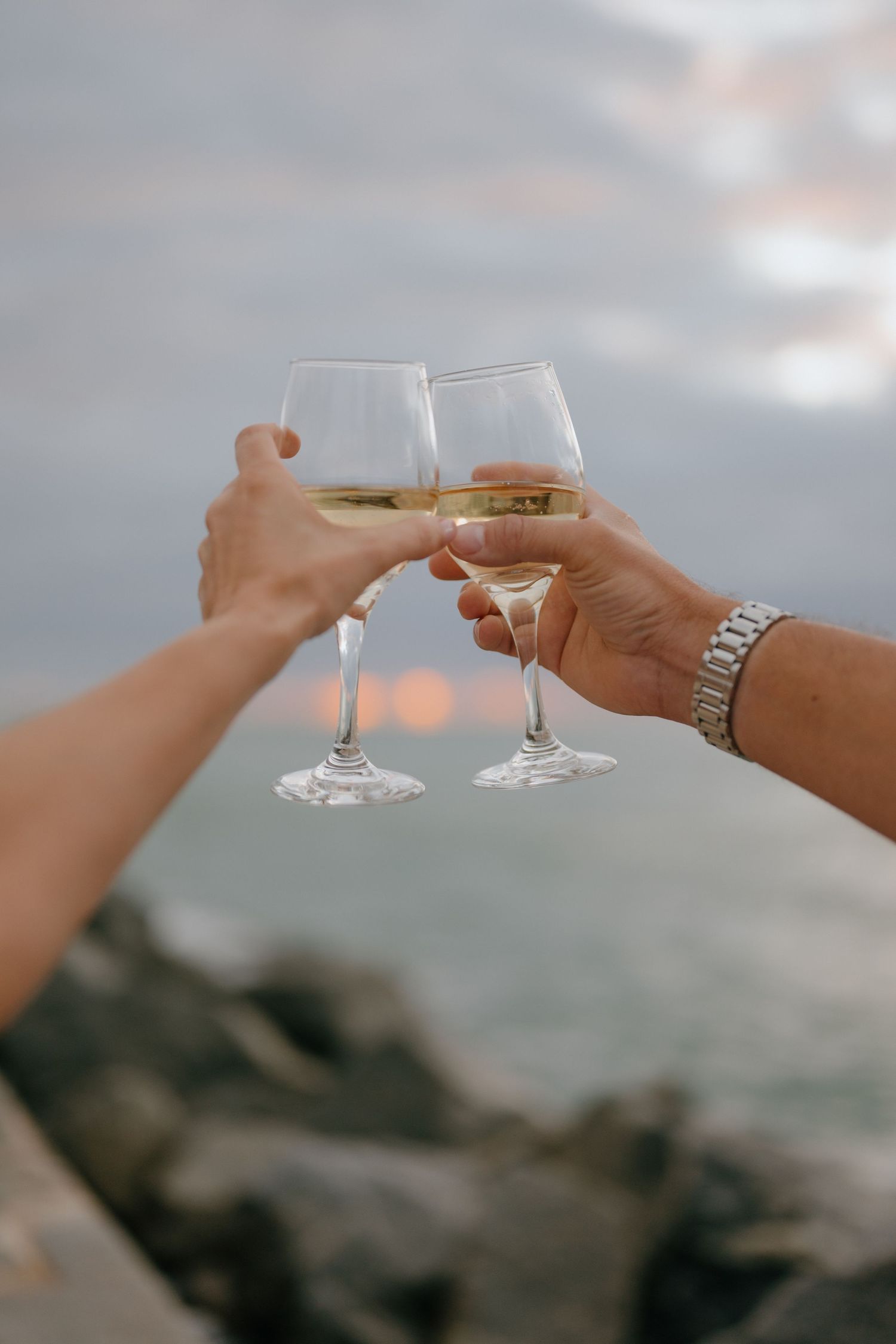 Two hands toasting with white wine glasses at sunset against a blurry rocky coastal background.