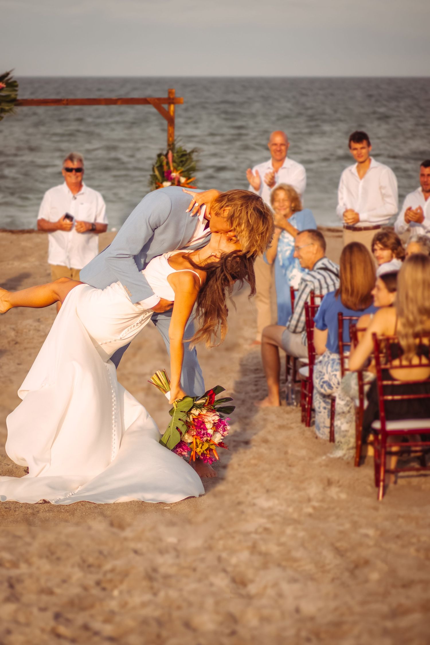 Bride and groom share a romantic dip kiss at a beach wedding ceremony with guests seated on sand.
