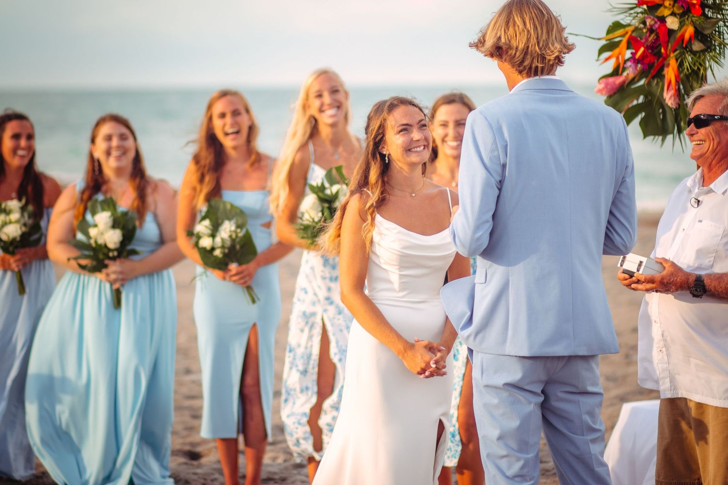 Bride smiling joyfully at groom during romantic beach wedding ceremony with bridesmaids in light blue dresses.
