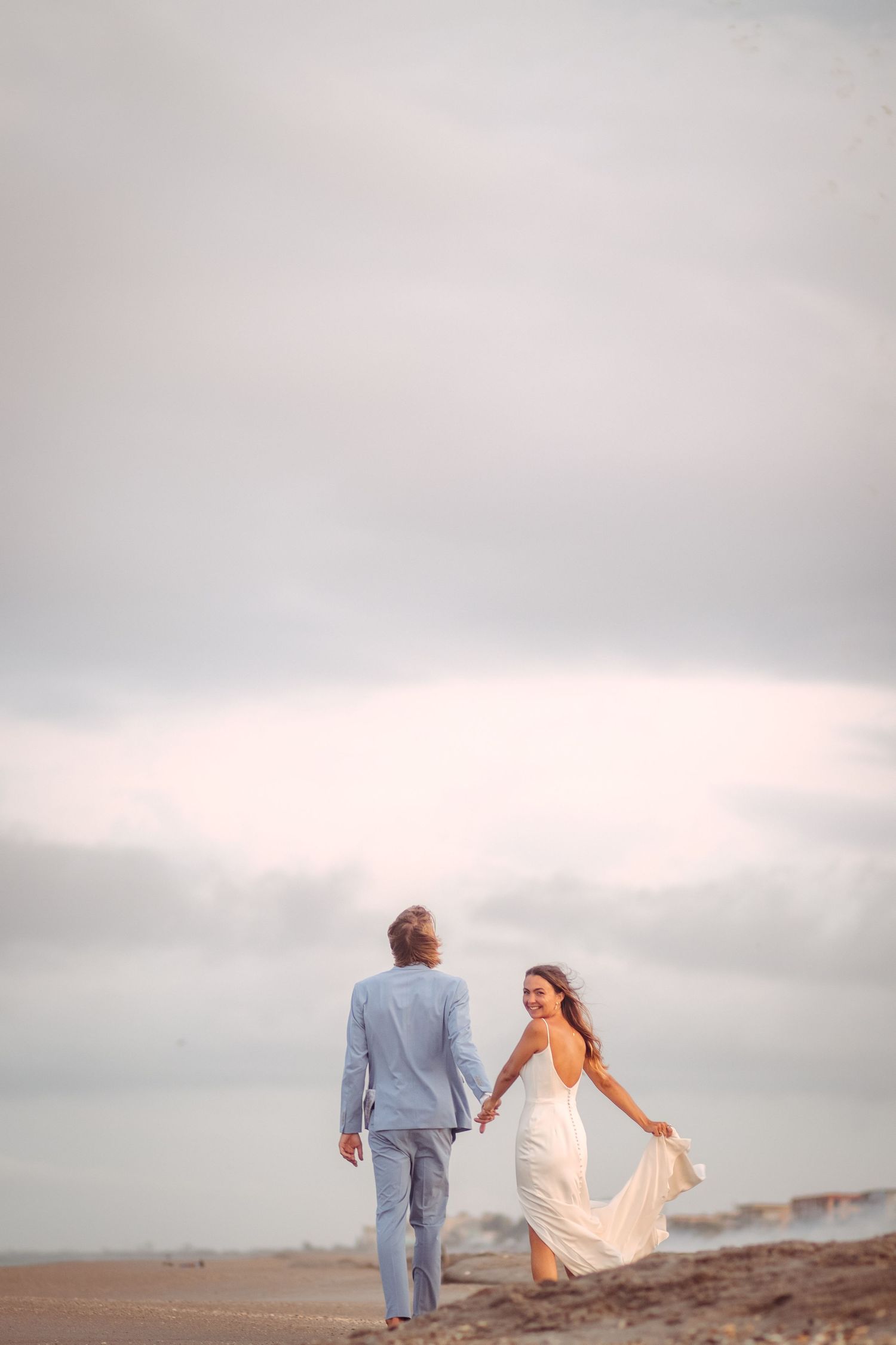 A couple in wedding attire walks hand-in-hand on a beach, the bride smiling back in a flowing white dress.