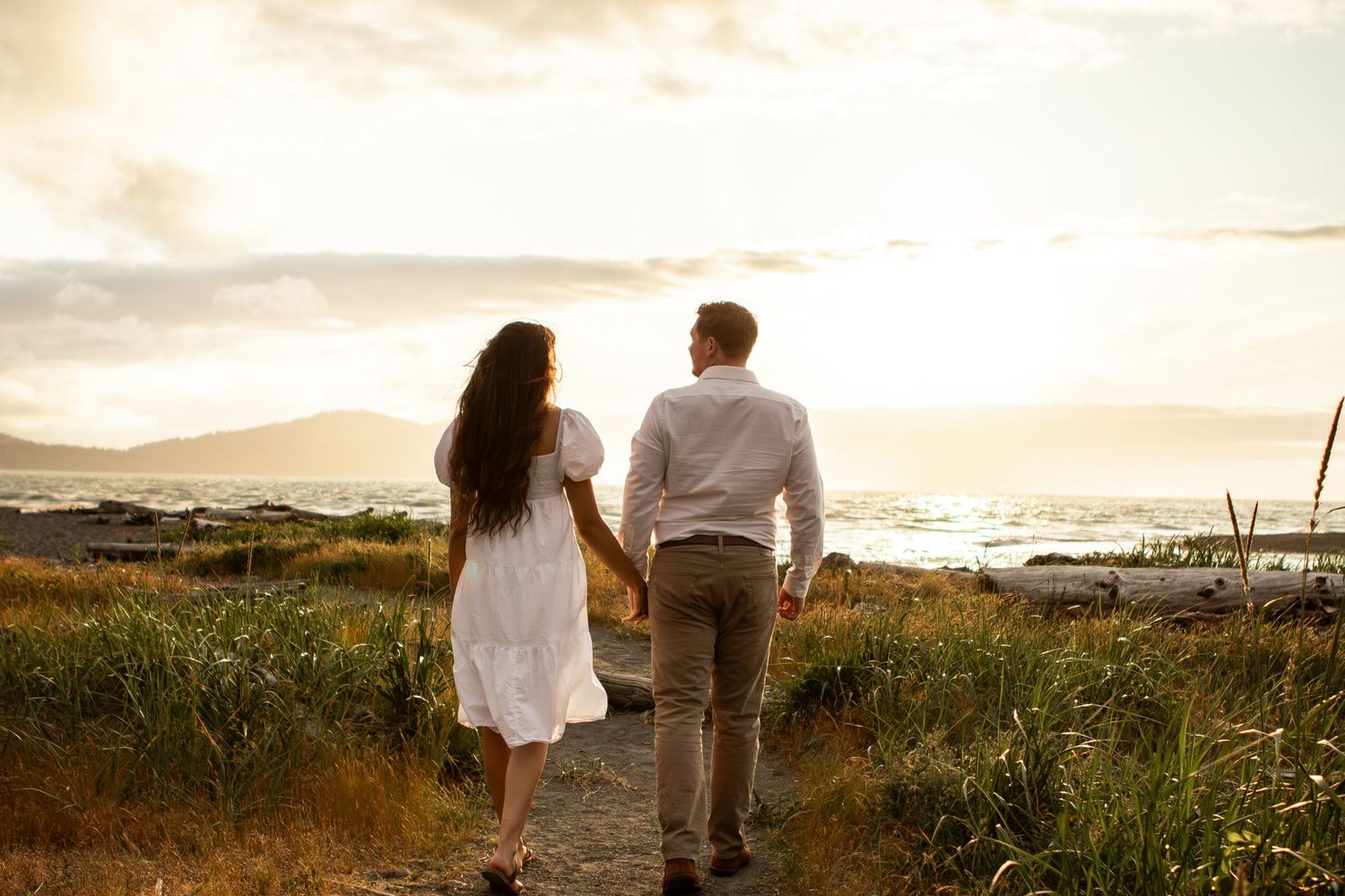 Couple walking along scenic coastal path during golden sunset with mountains in background.