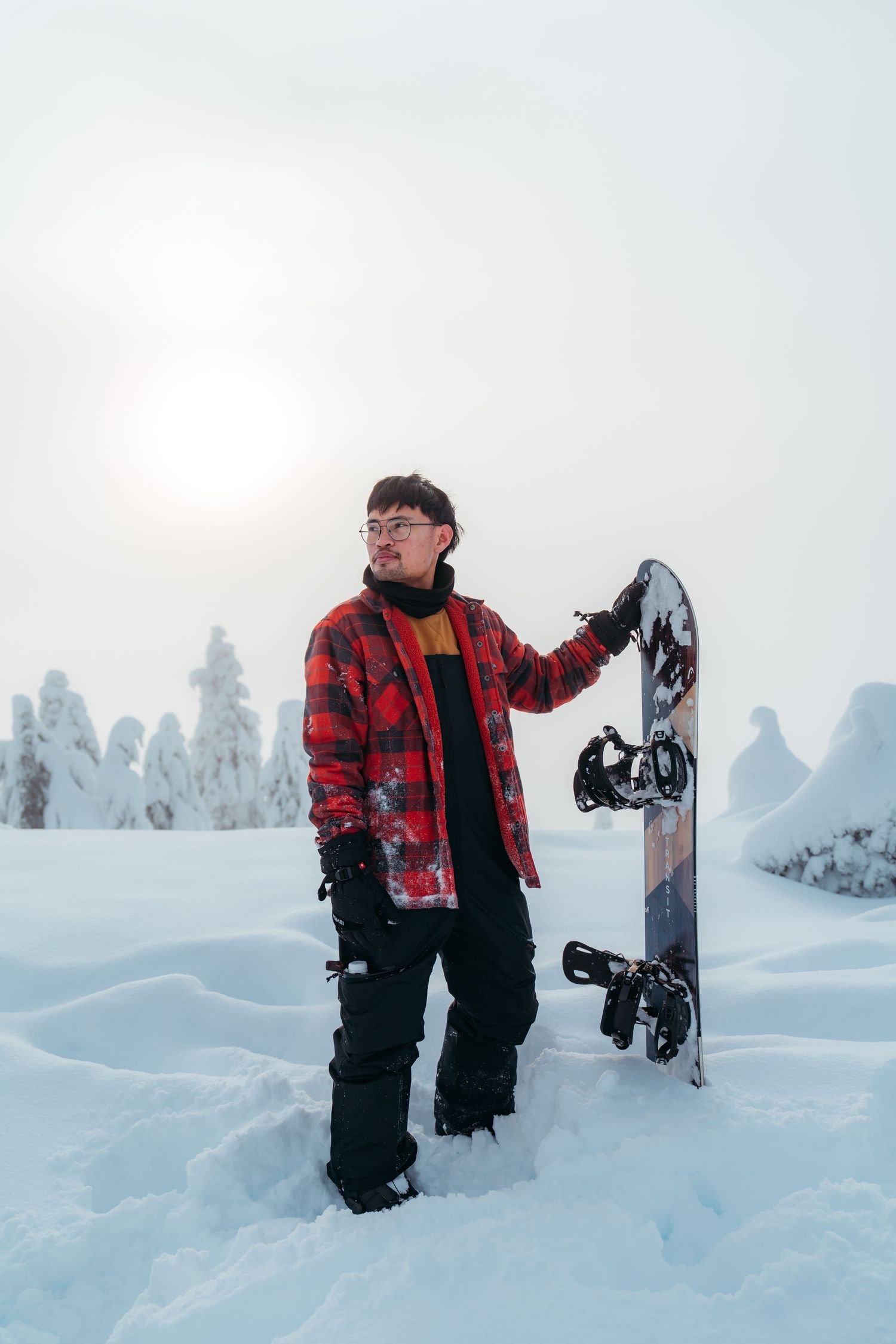 Winter sports enthusiast in red plaid jacket holding snowboard in snowy forest setting.