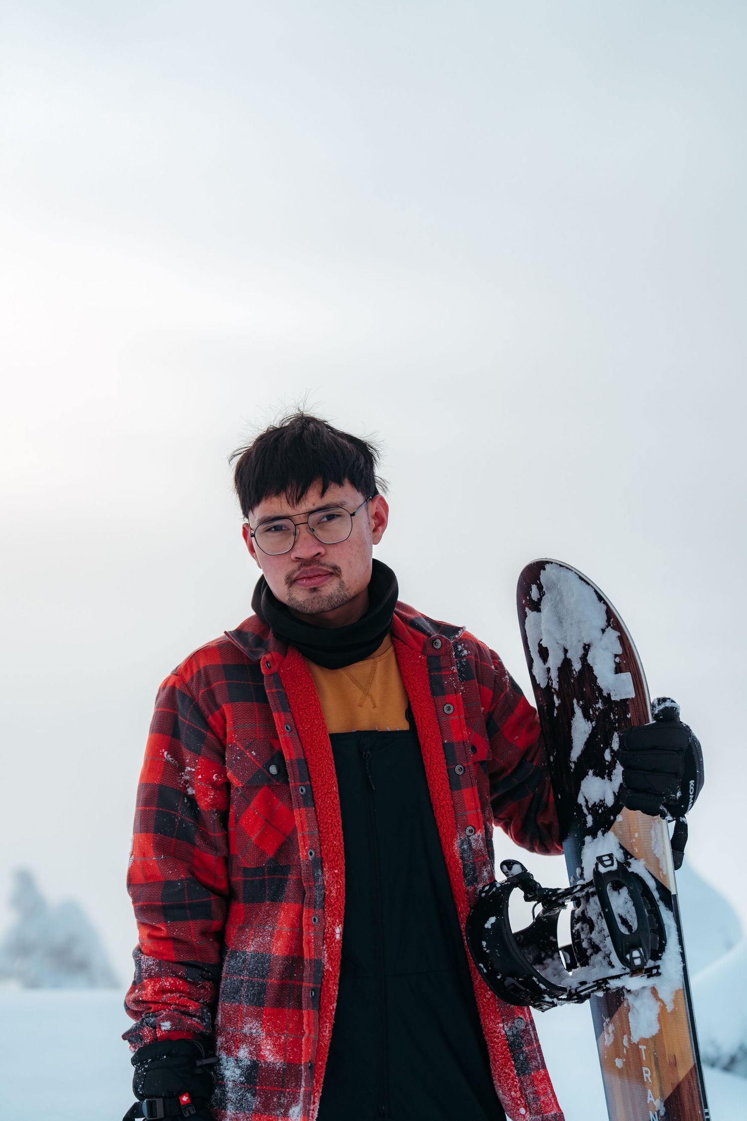 Winter sports enthusiast in red plaid jacket holding snowboard against snowy background.