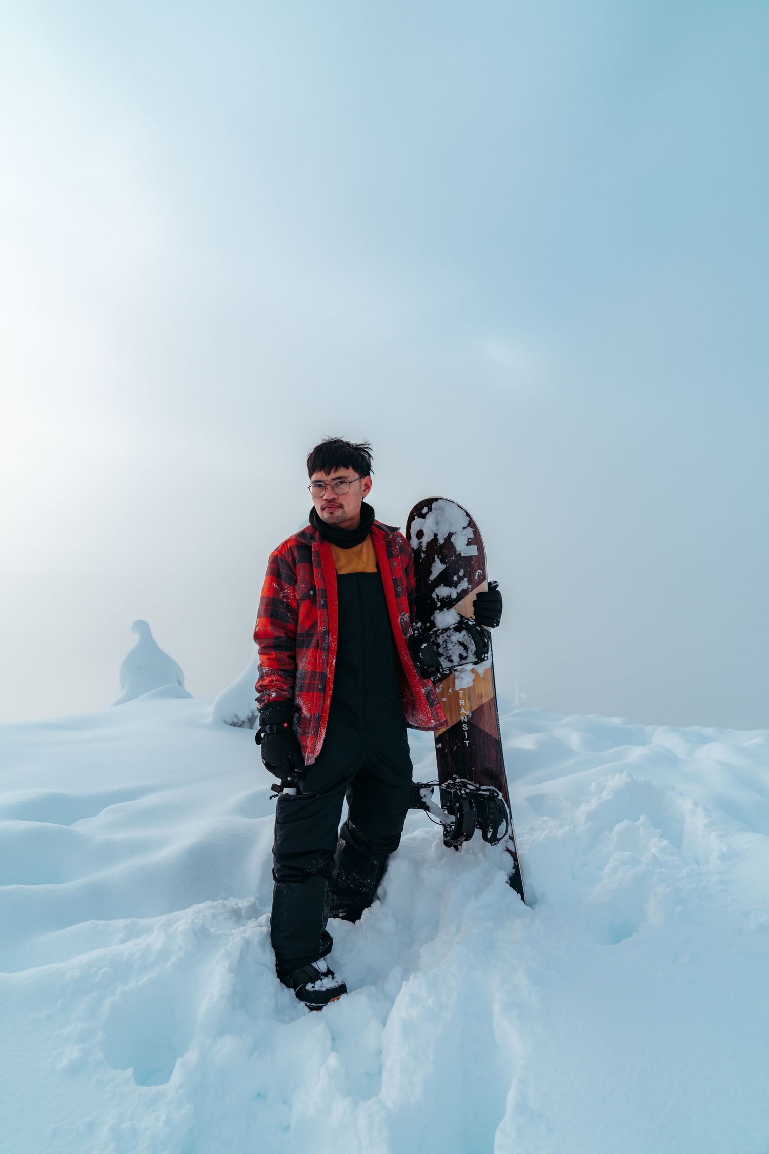 Person in red plaid jacket posing with snowboard in snowy winter landscape.