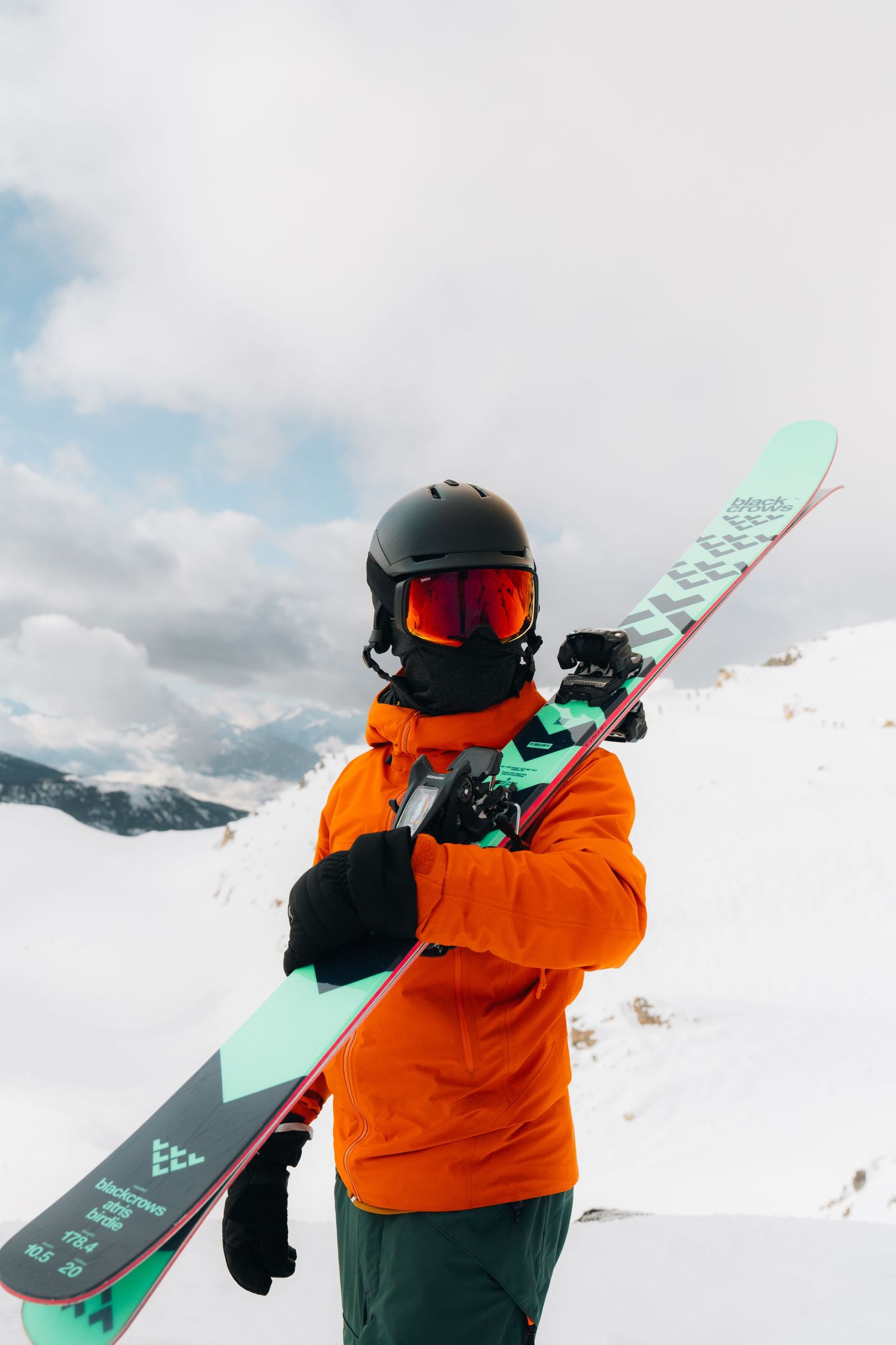 Skier in bright orange jacket holds teal skis against snowy mountain backdrop with cloudy sky.
