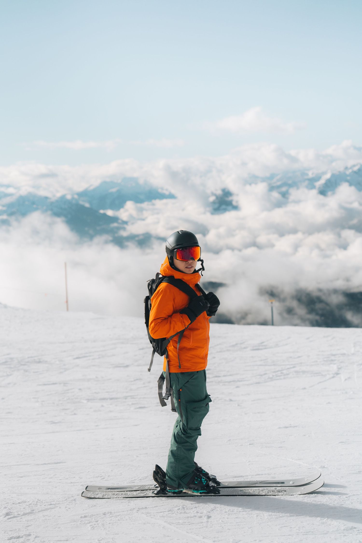 Skier in orange jacket and black helmet standing on snowy slope above cloud-covered mountains.