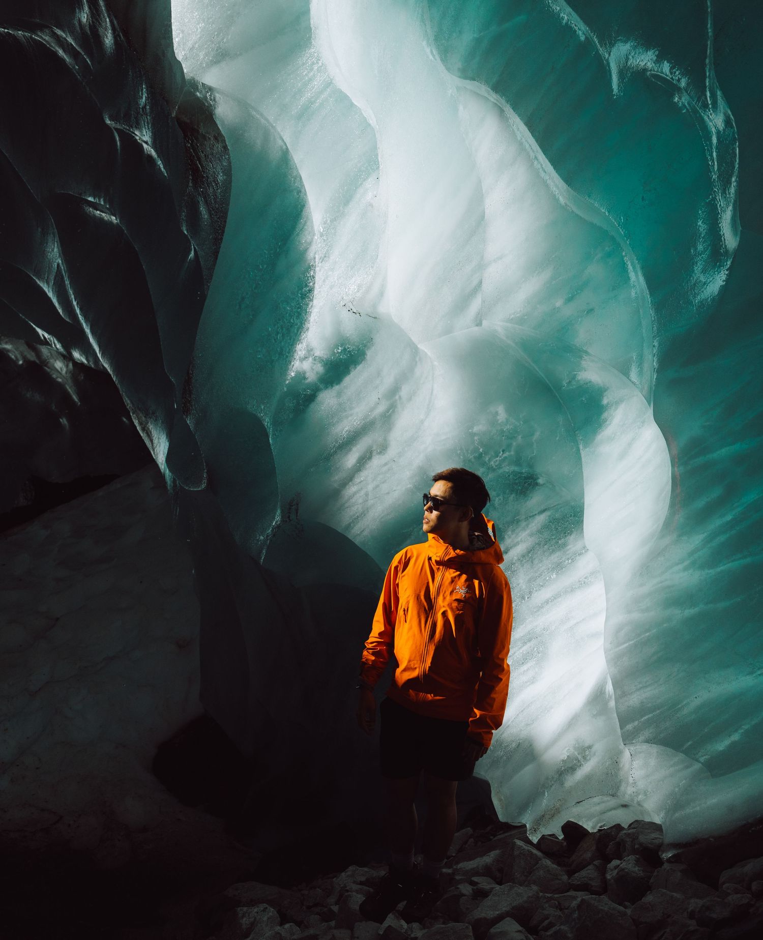 Explorer in orange jacket stands inside turquoise ice cave with dramatic glacial formations.