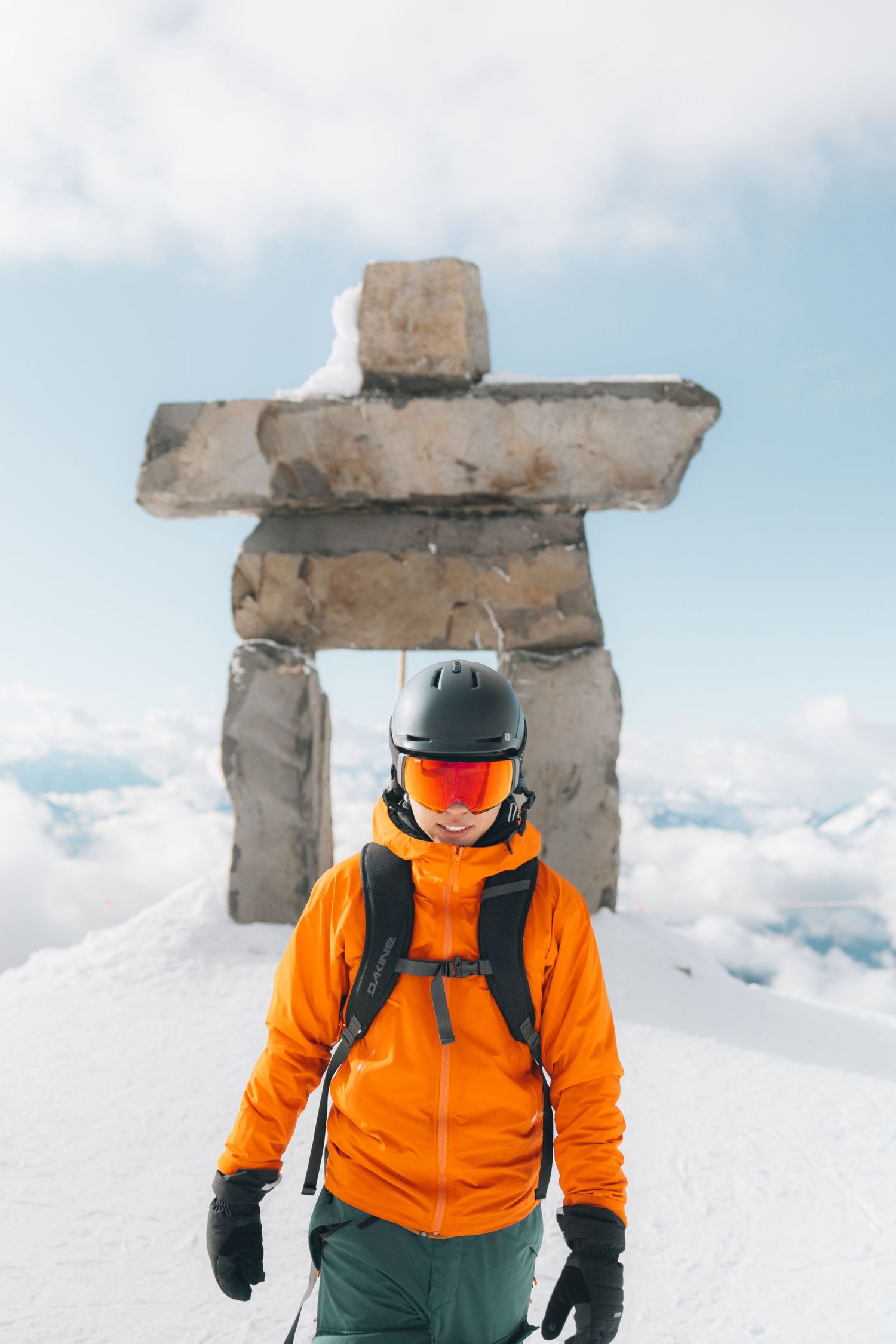 Skier poses beneath traditional Inukshuk stone monument on snowy mountain peak.