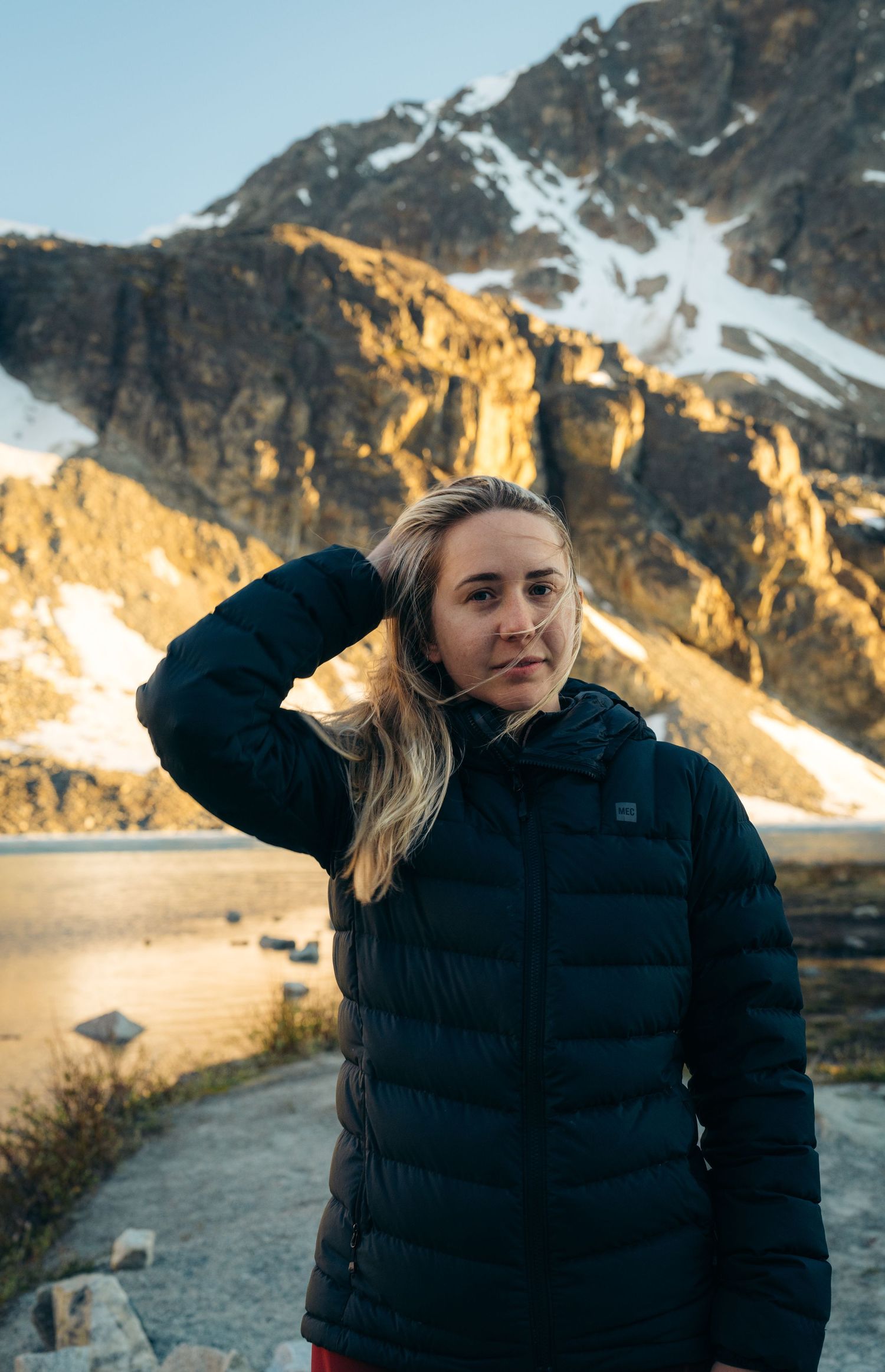 Person in black winter jacket against sunlit mountain backdrop shields eyes from sun.
