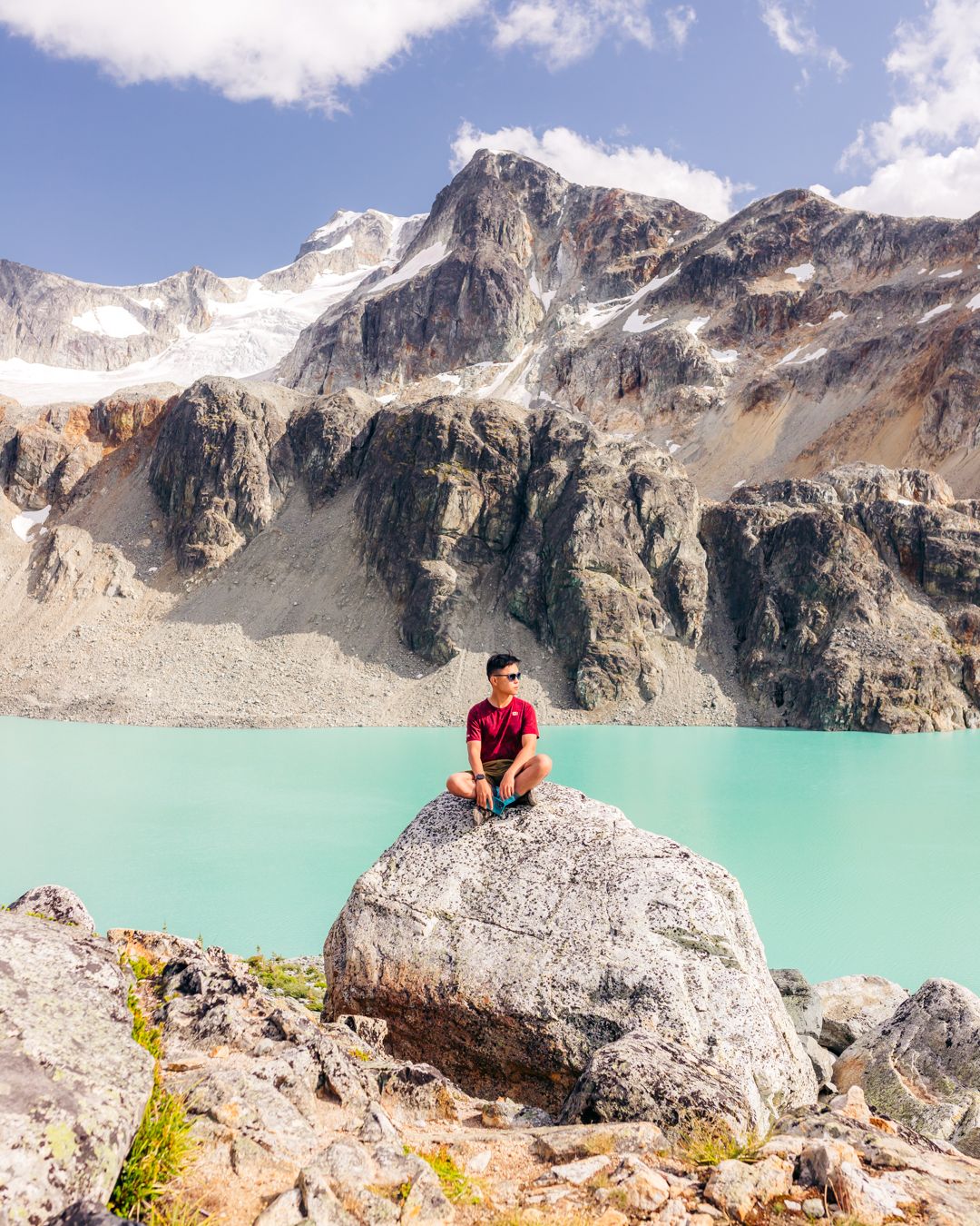 Figure sitting on large boulder overlooking turquoise alpine lake surrounded by snow-capped mountains.