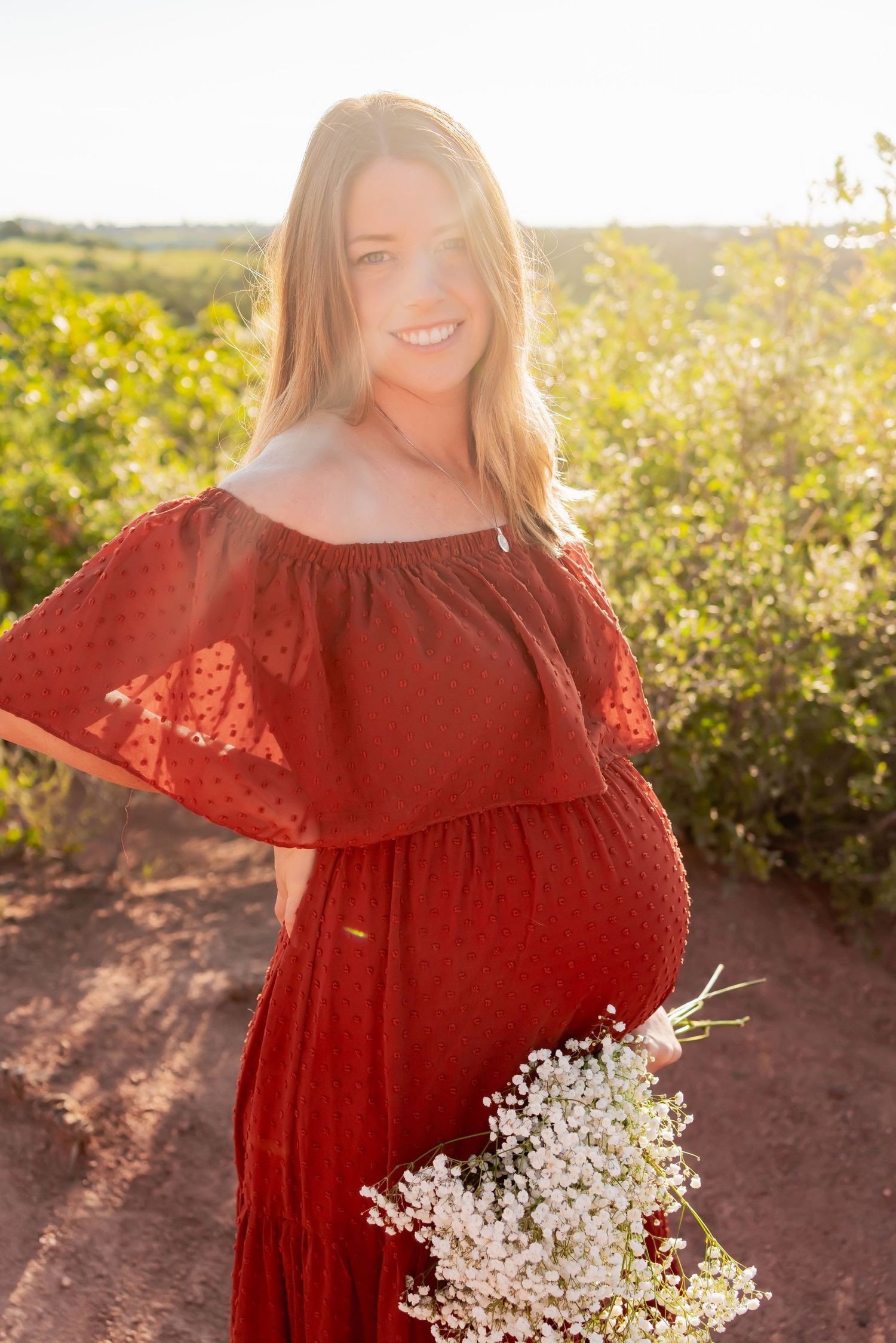 Sun-drenched maternity portrait in red lace dress holding baby's breath flowers.