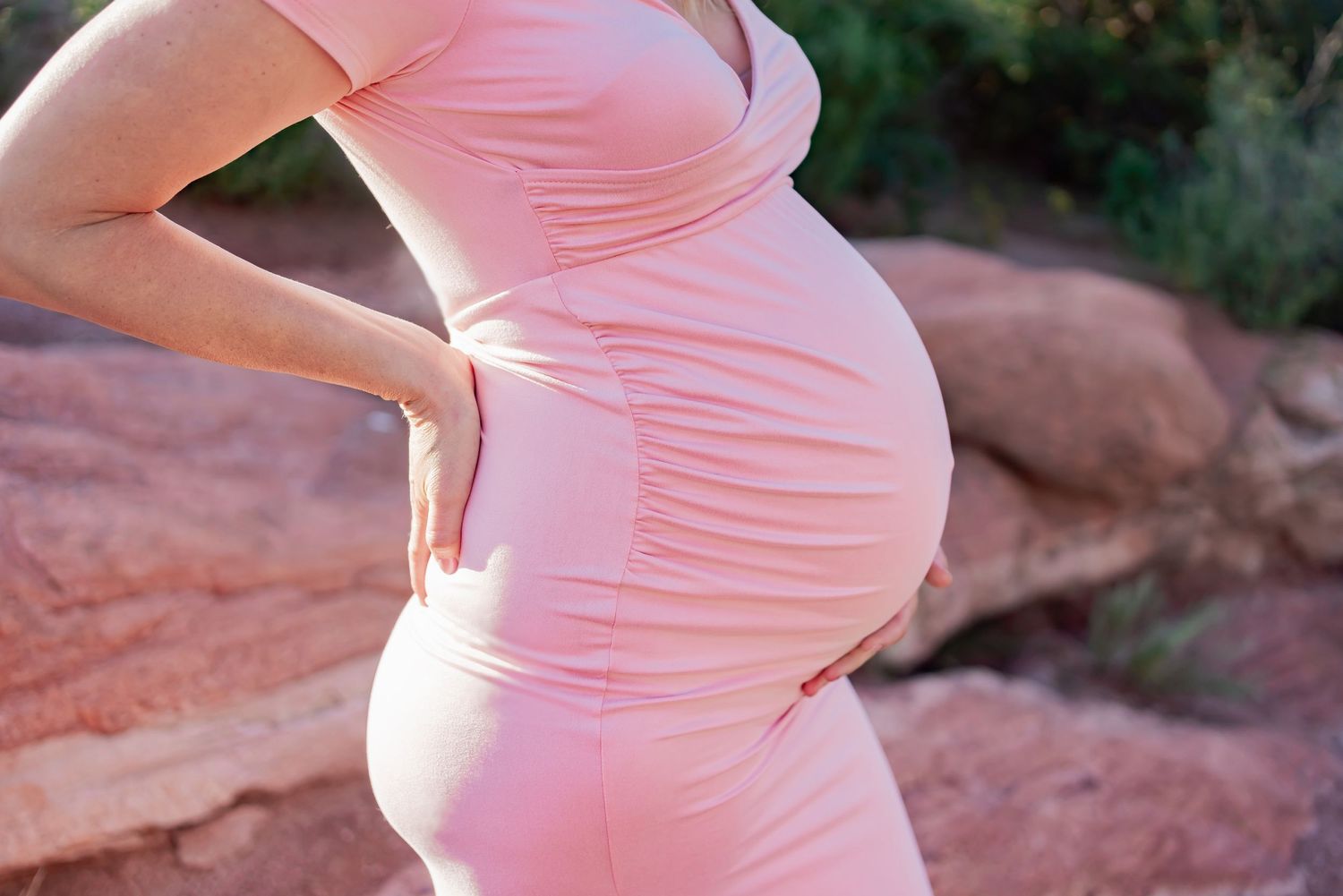 A side view of an expectant mother in a pink dress highlighting her baby bump against a red rock backdrop.
