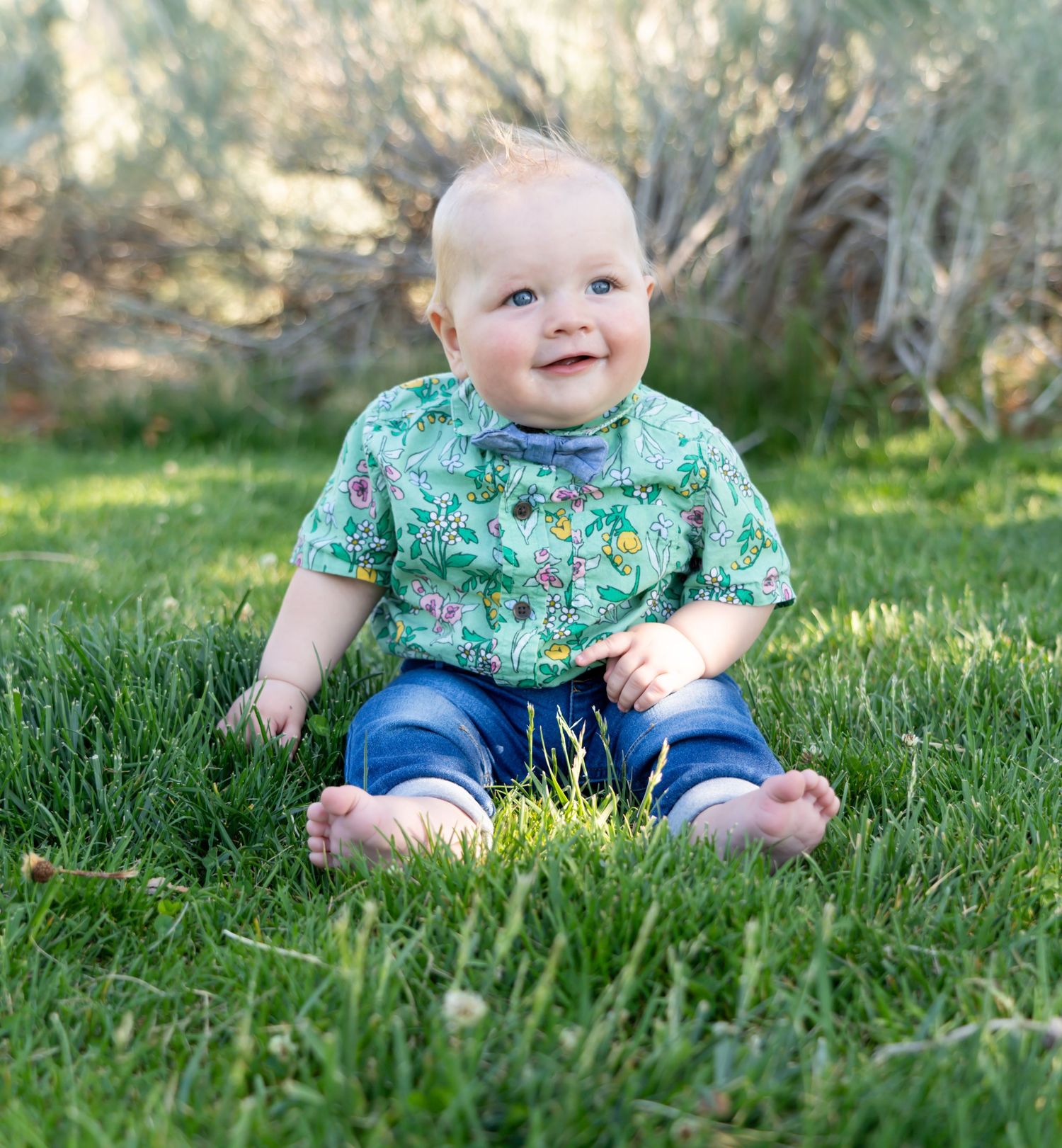 Smiling baby in tropical print shirt sitting on grass in garden setting.