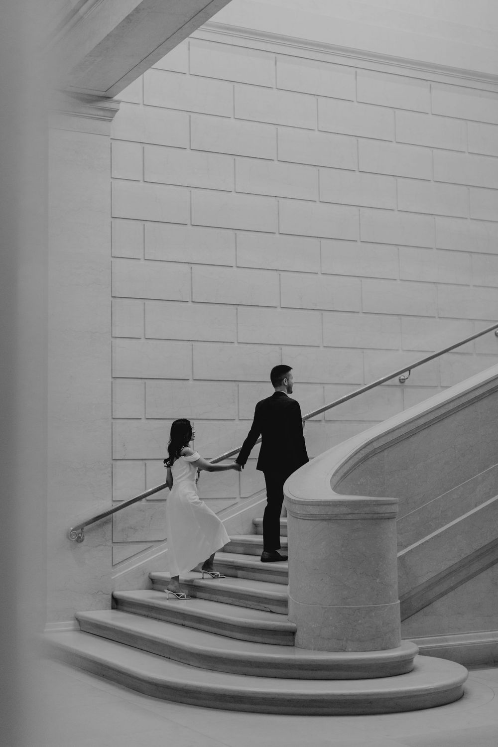 Black and white photo of bride and groom holding hands walking up a sweeping marble staircase.