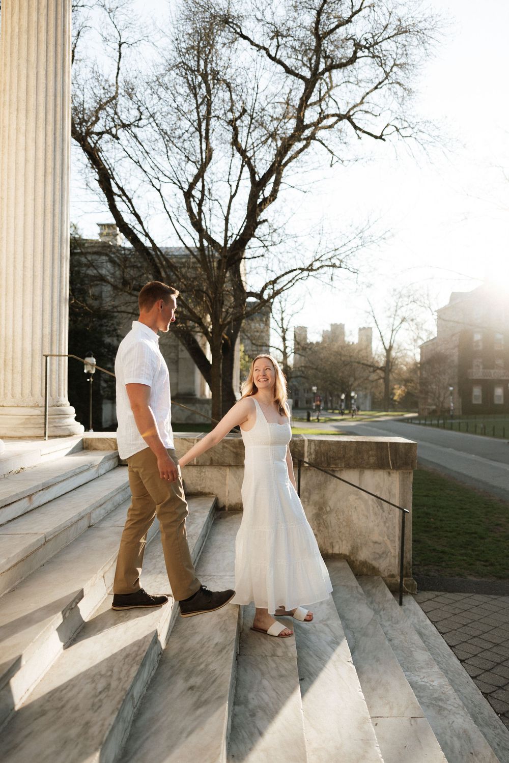 Couple holding hands on sunlit stone steps, woman in white dress, man in white shirt and khakis, bare trees in background.