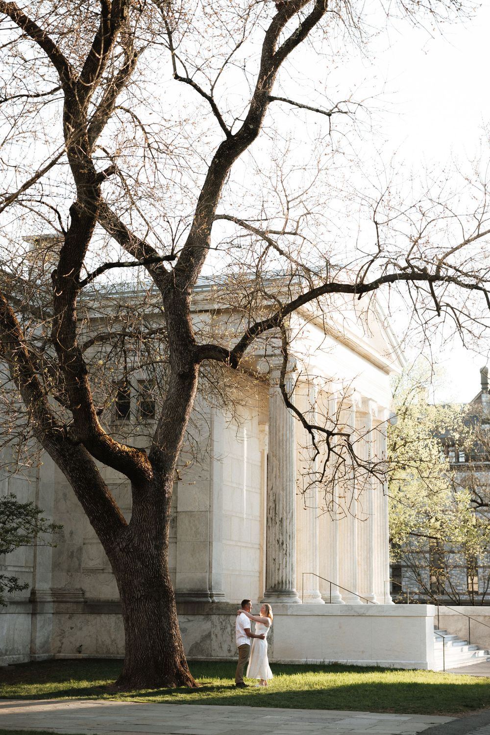 Couple embracing under a bare tree beside a grand white neoclassical building during a romantic outdoor wedding photo session.