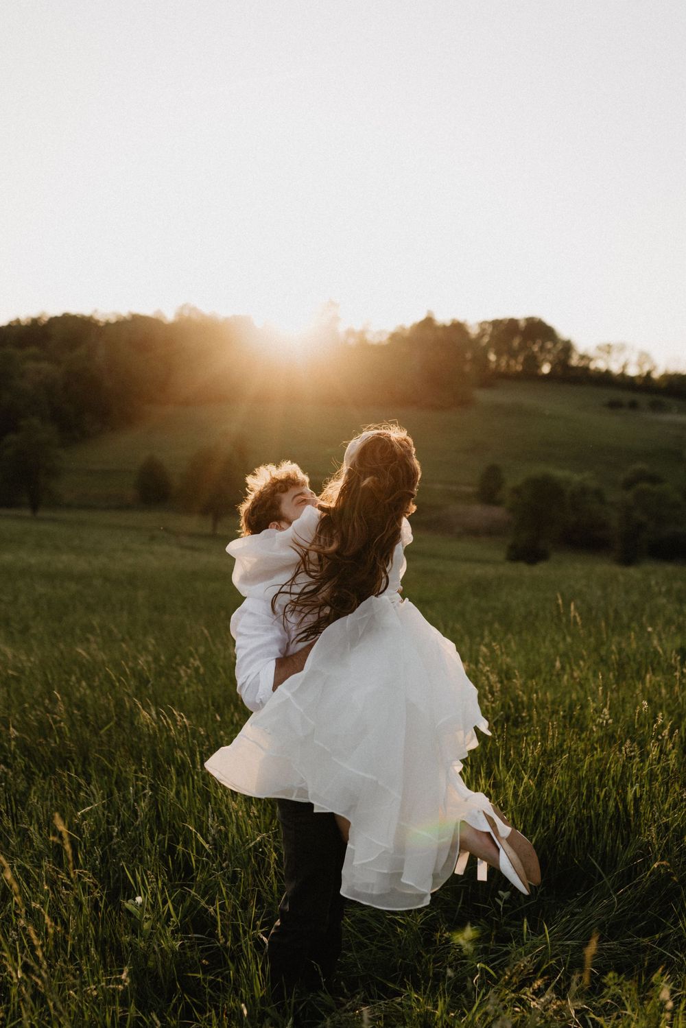 Groom lifting bride in white dress at golden sunset in a lush green countryside field.
