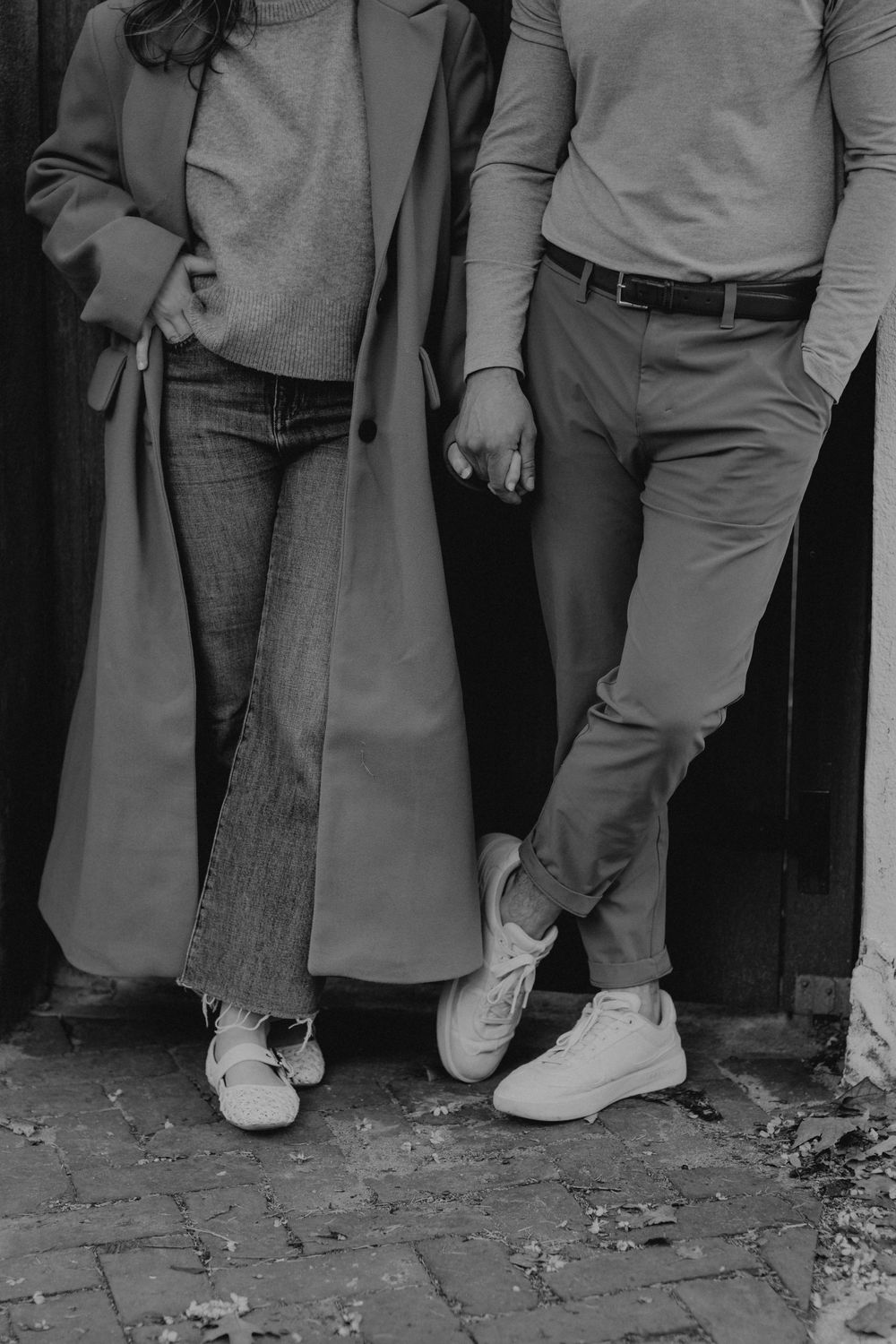 Black and white photo of a couple holding hands, wearing casual outfits and white sneakers on a cobblestone street.