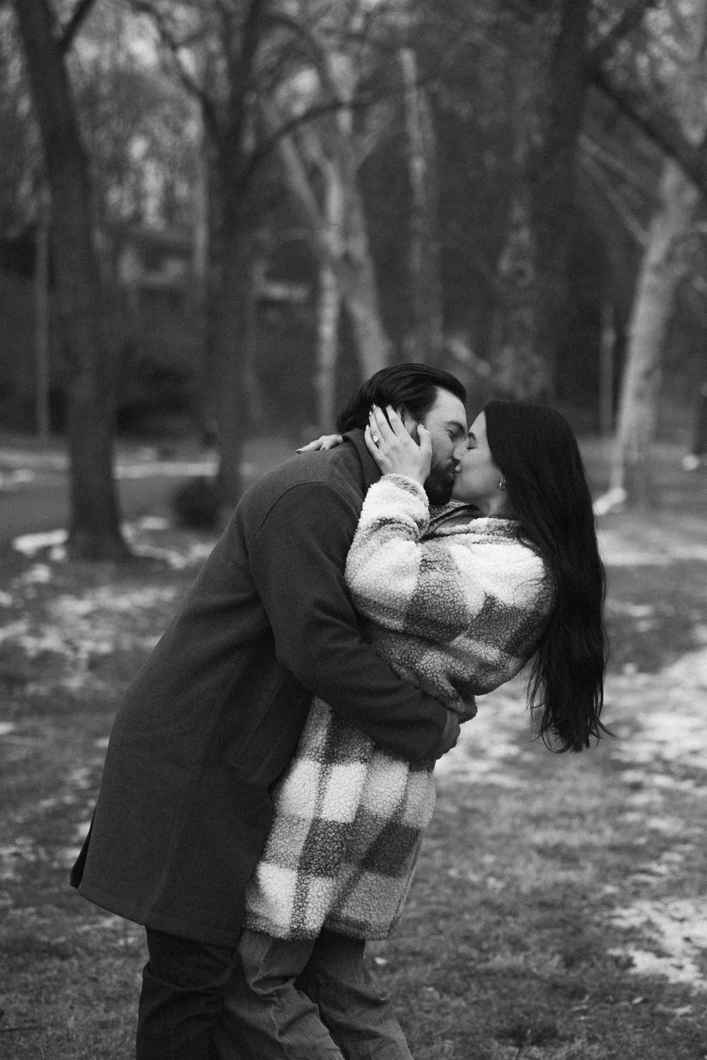 Black and white photo of couple sharing a passionate kiss among bare winter trees.