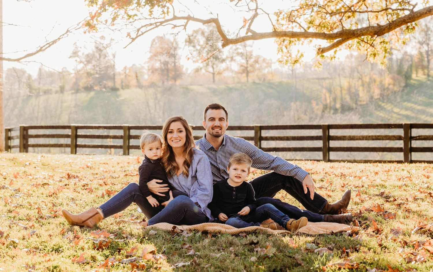 Family lounging together on fallen autumn leaves with a fence in background.