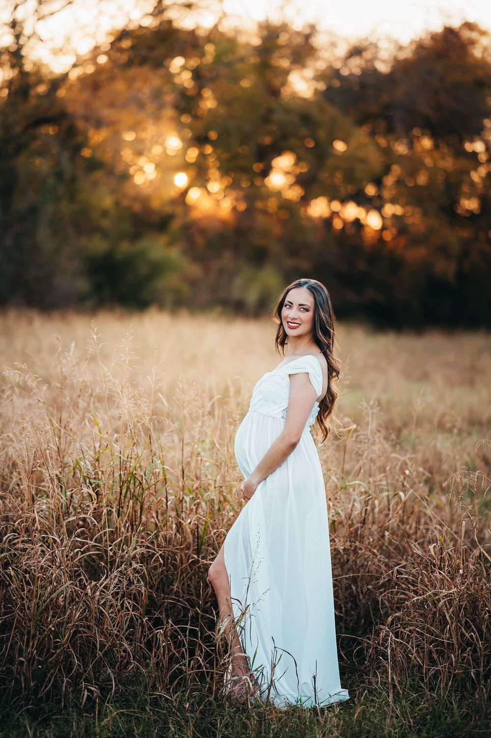 Pregnant woman in white off-shoulder gown standing in golden field at sunset during maternity photoshoot.