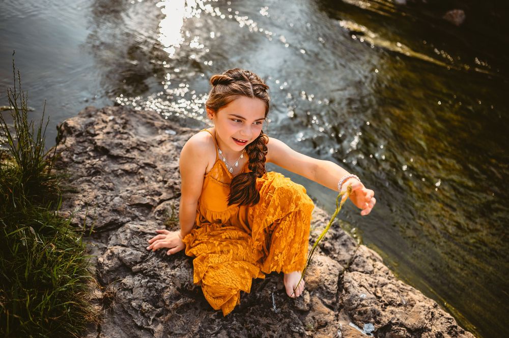 A young girl in a yellow outfit sitting on rocks beside a sparkling river outdoors.