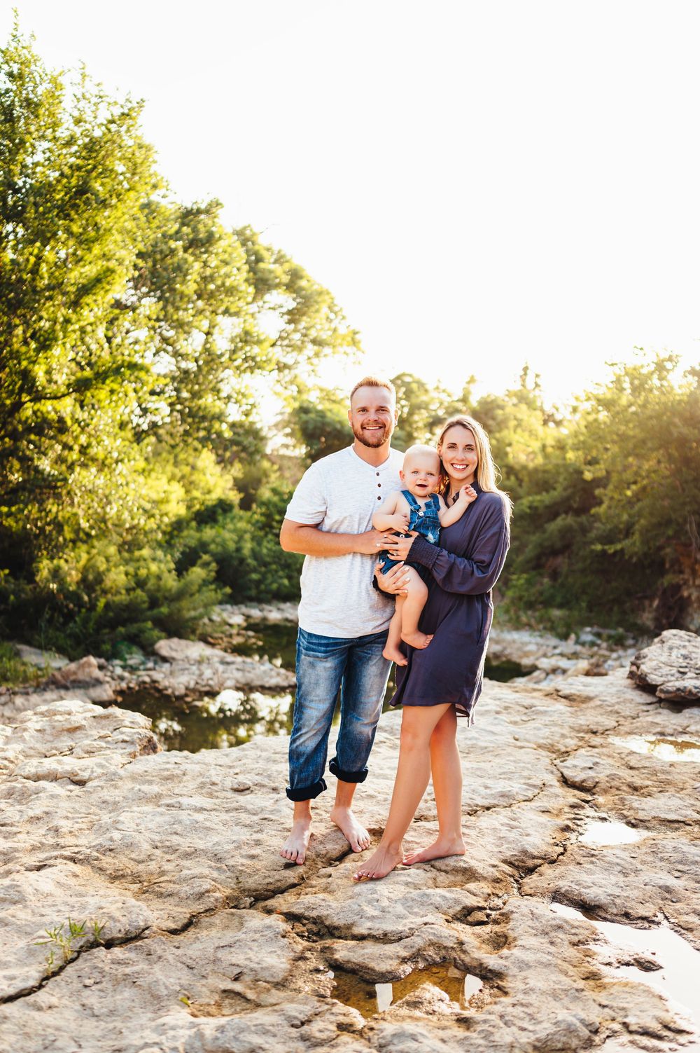 Young family with baby posing on rocky riverbank surrounded by lush green trees in golden sunlight.