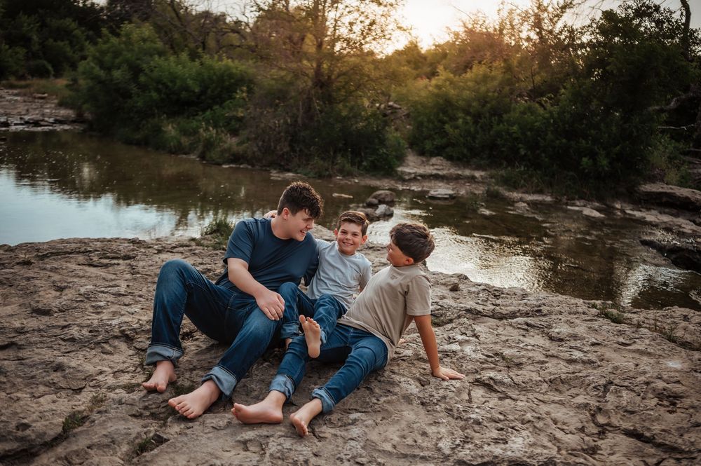 Three boys sitting barefoot on rocky ground near a river during a golden sunset.