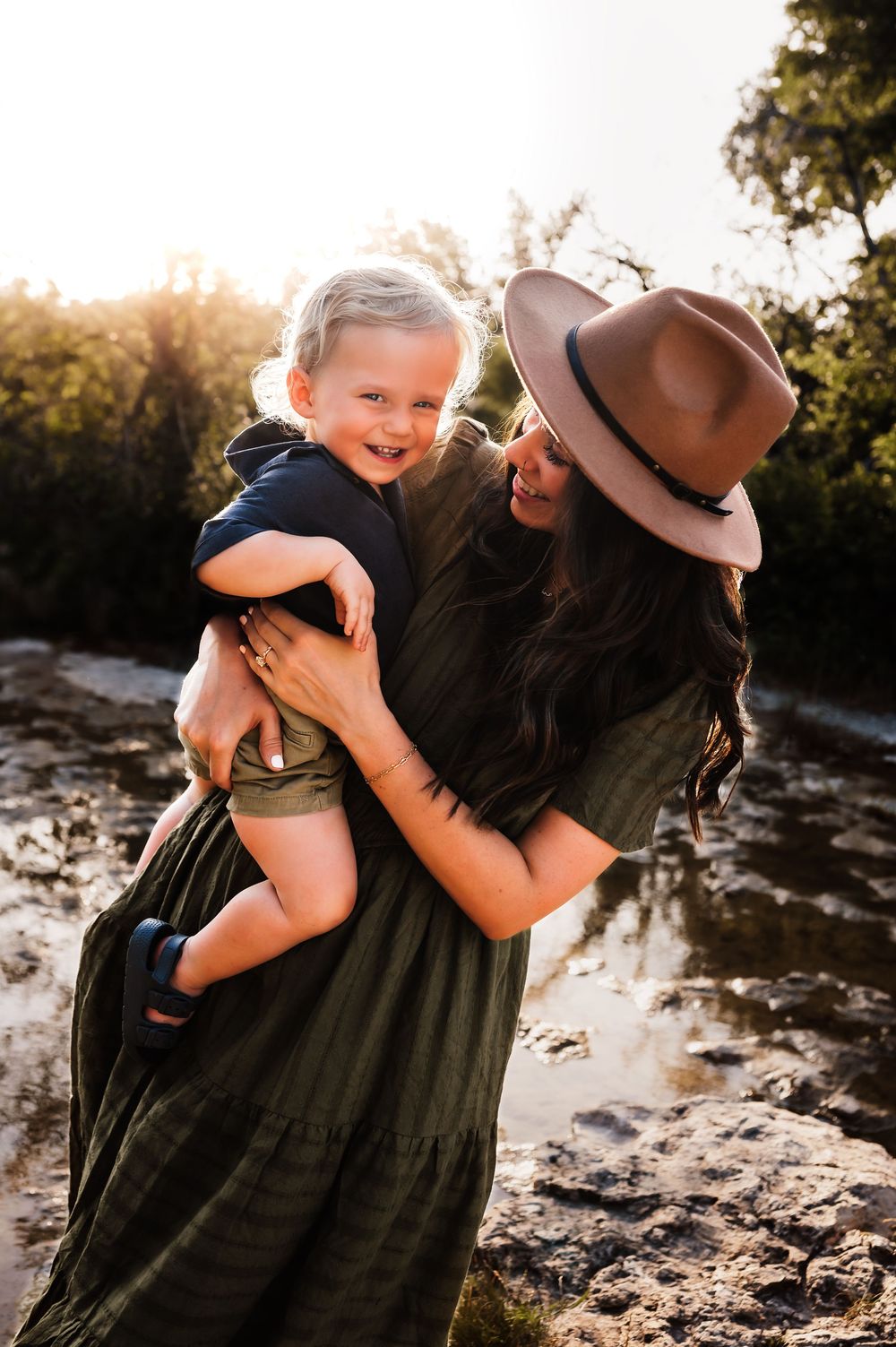 A smiling mother in a wide-brim hat holds her laughing toddler outdoors in golden sunlight.
