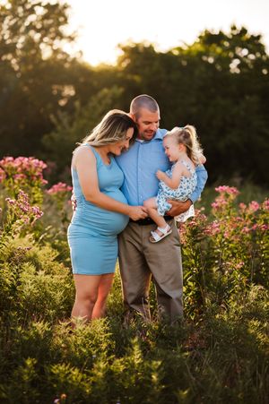 Pregnant couple in blue outfits holding toddler girl among pink wildflowers at golden hour.