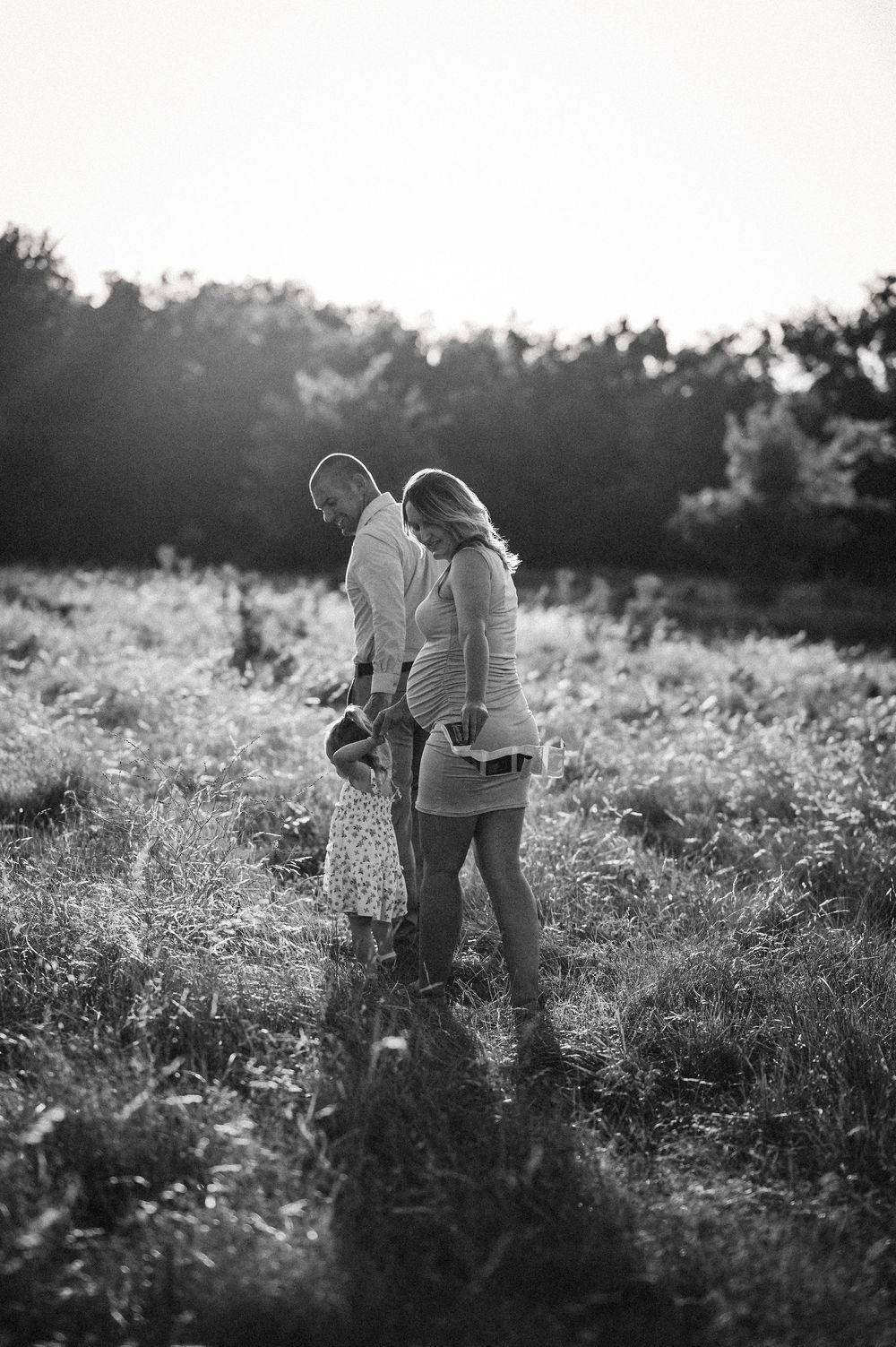 Black and white photo of pregnant couple and child walking through sunlit meadow at sunset.