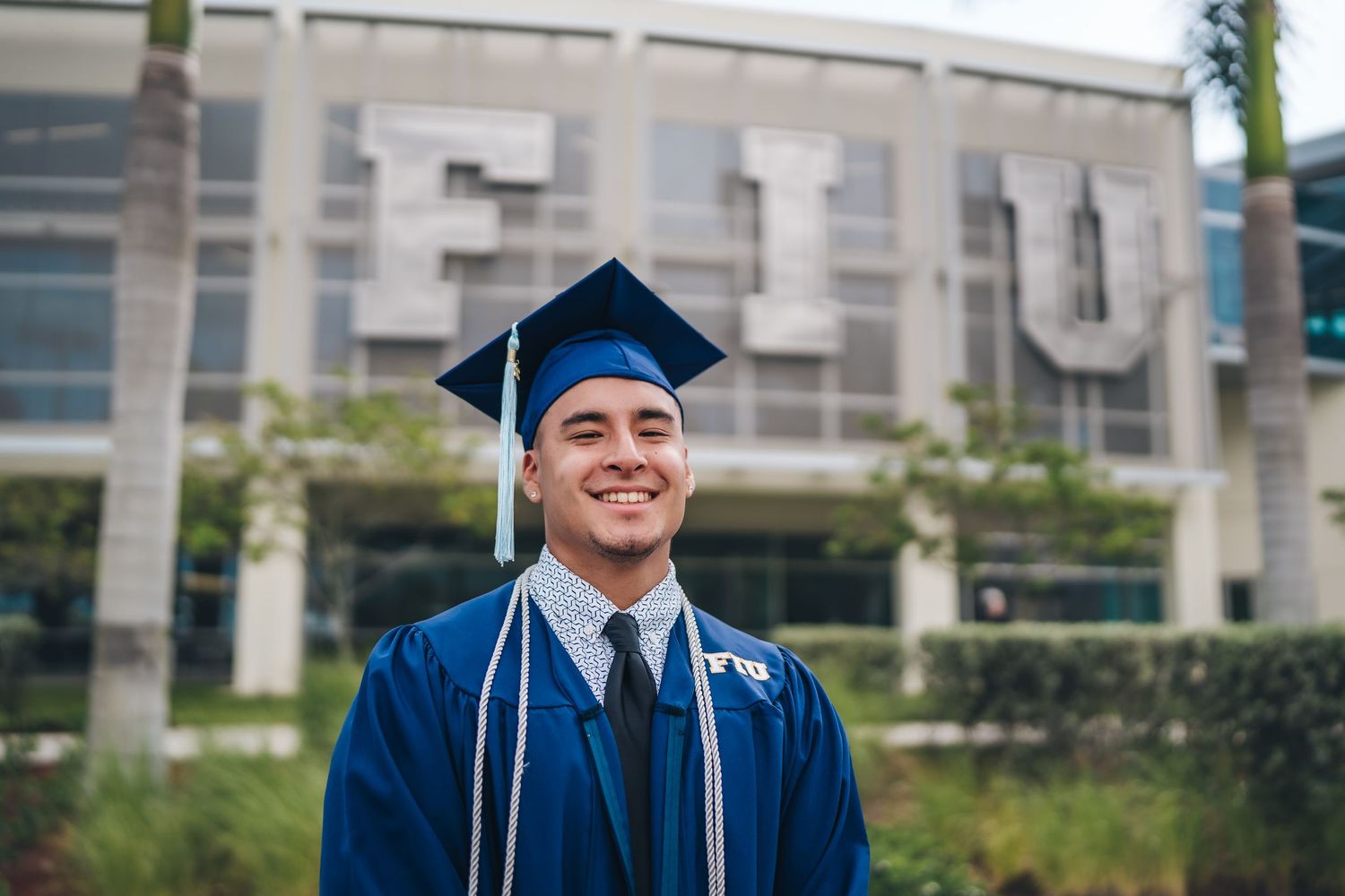 Graduate in blue cap and gown stands proudly in front of university building.