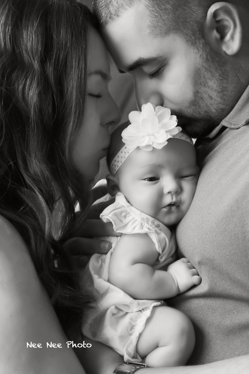 Black and white photo of parents tenderly kissing their newborn baby girl wearing a floral headband. Embracing Life’s Beauty