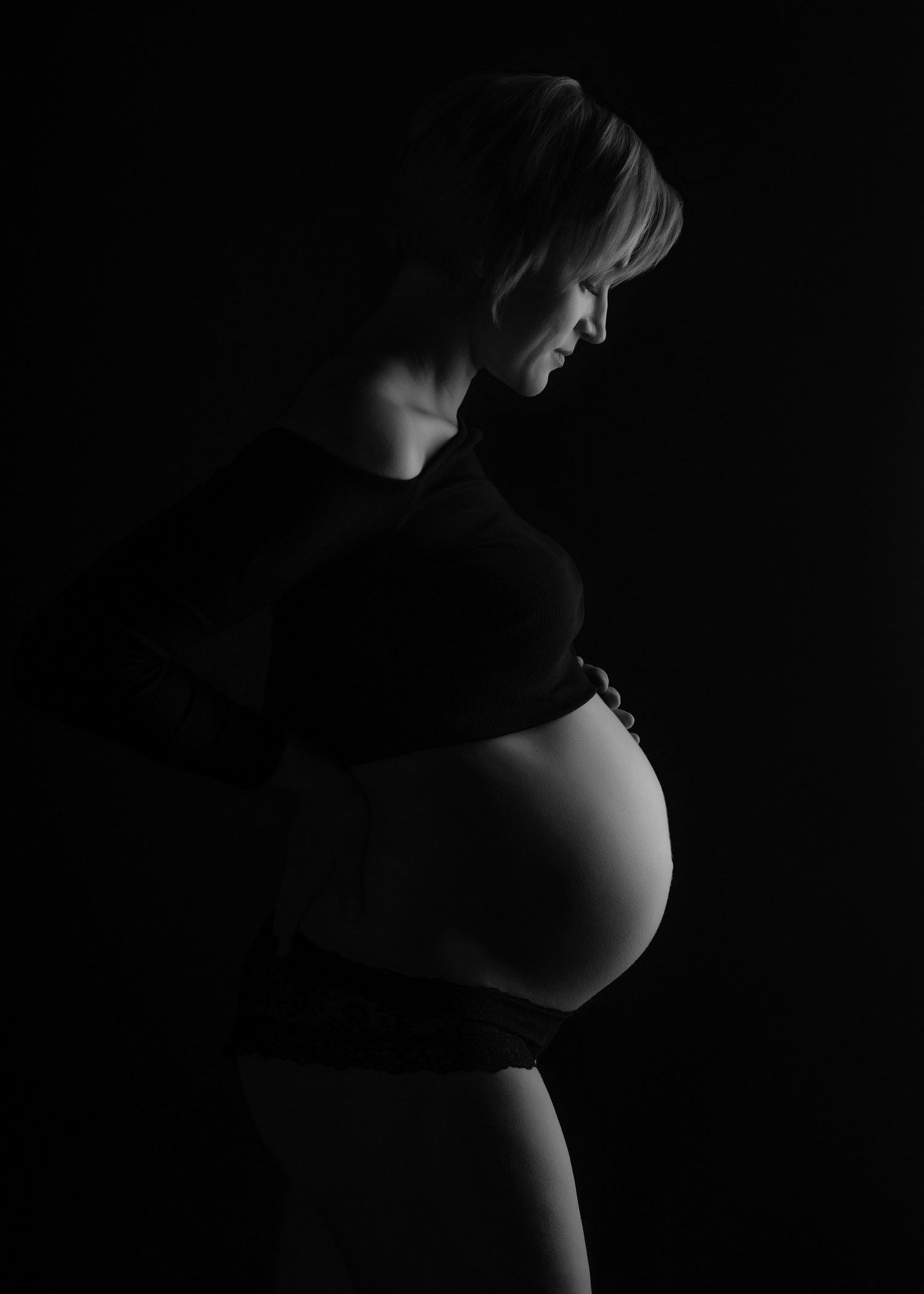 a black and white photo in studio of a pregnant woman softly smiling down at her baby bump for a maternity photo shoot
