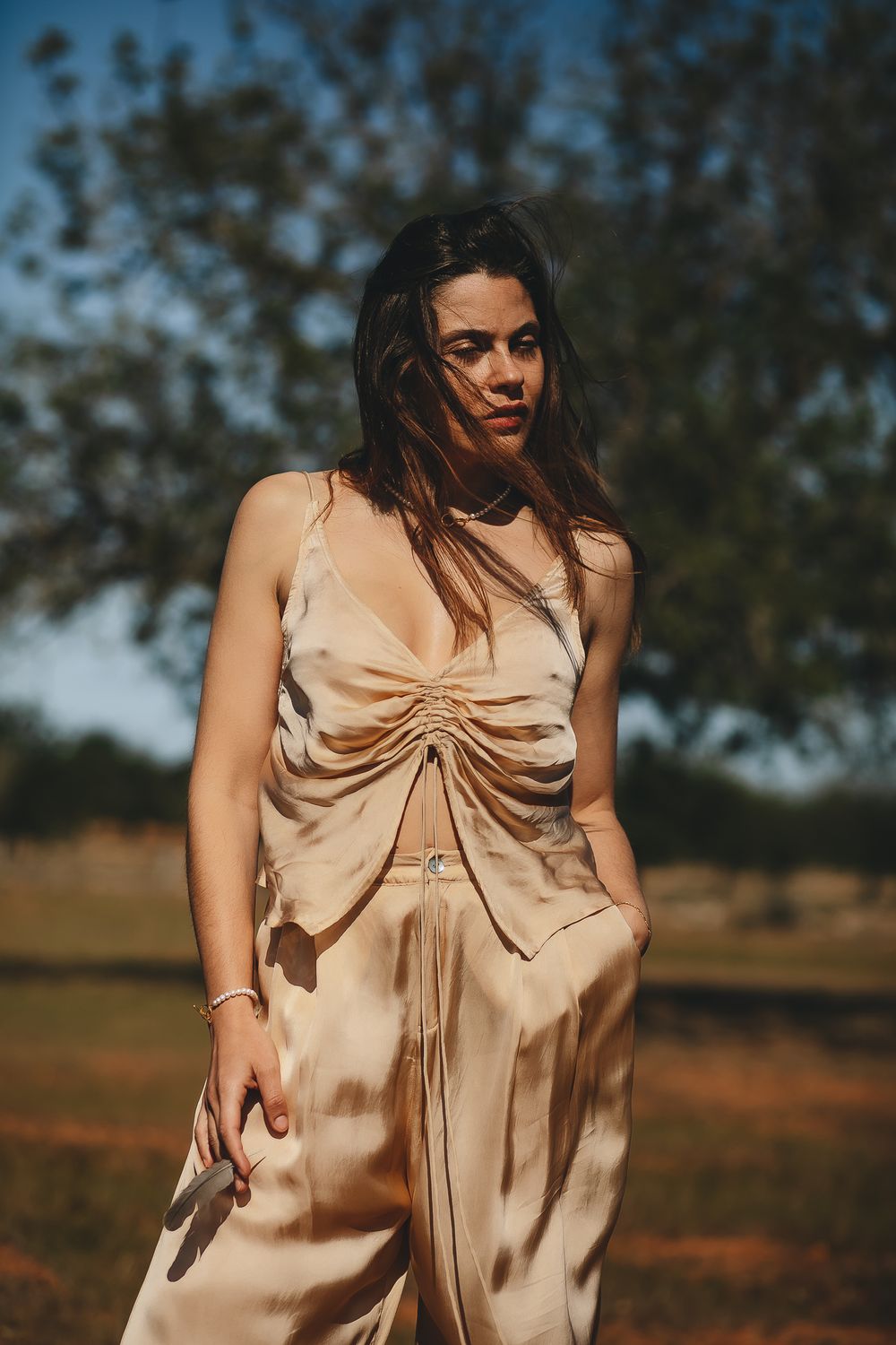 Woman in beige dress posing in a sunny field with trees in background.