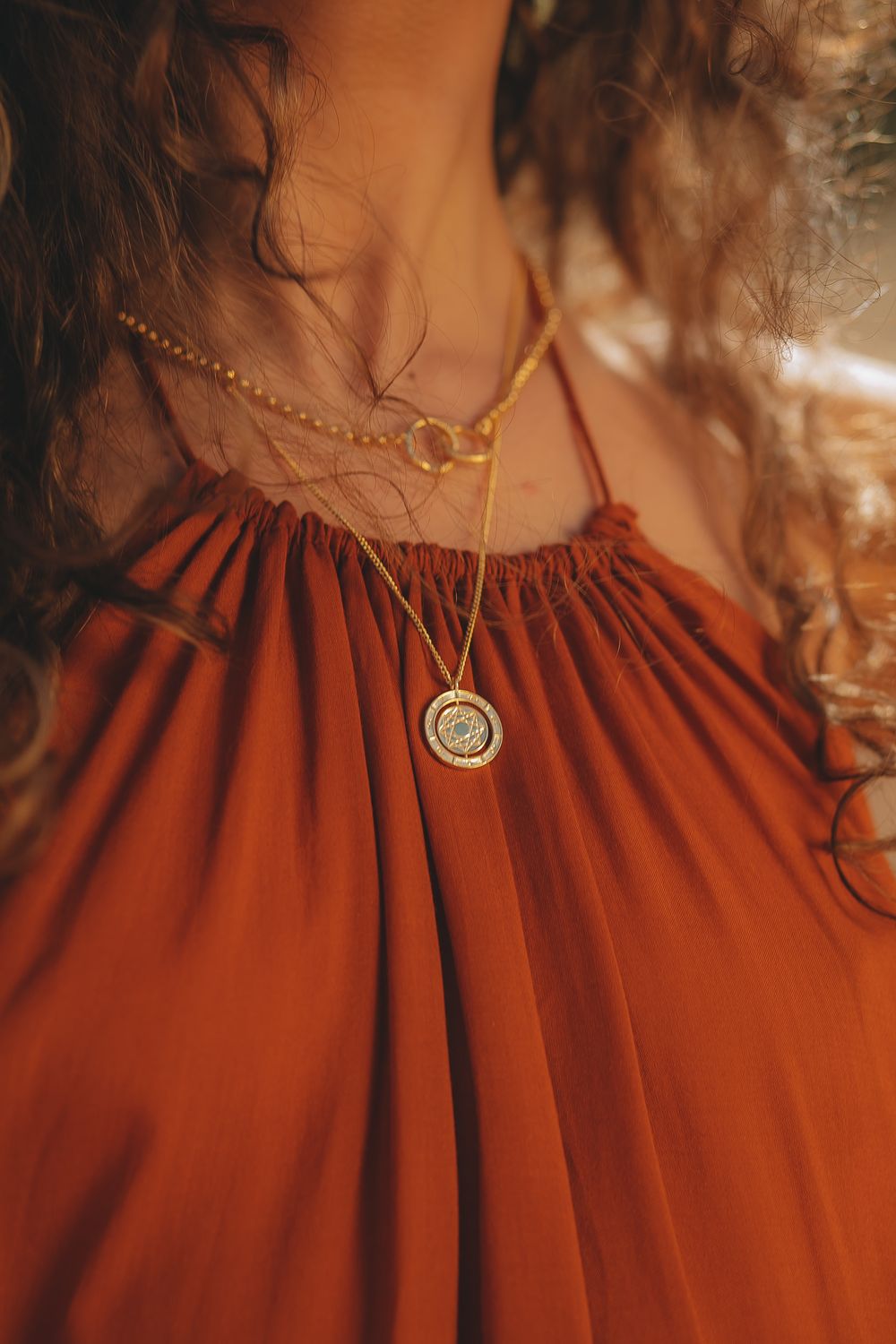 Close-up of a delicate gold necklace with pendant against a rust-colored gathered fabric dress.