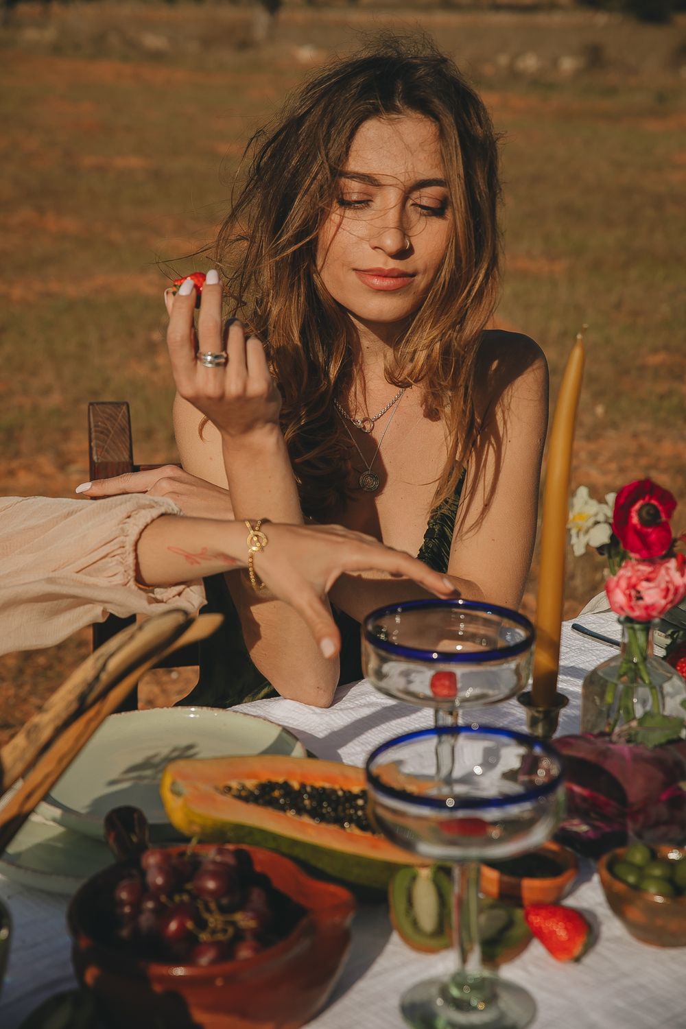 Outdoor picnic scene with fresh fruit and flowers on white tablecloth in golden sunlight.